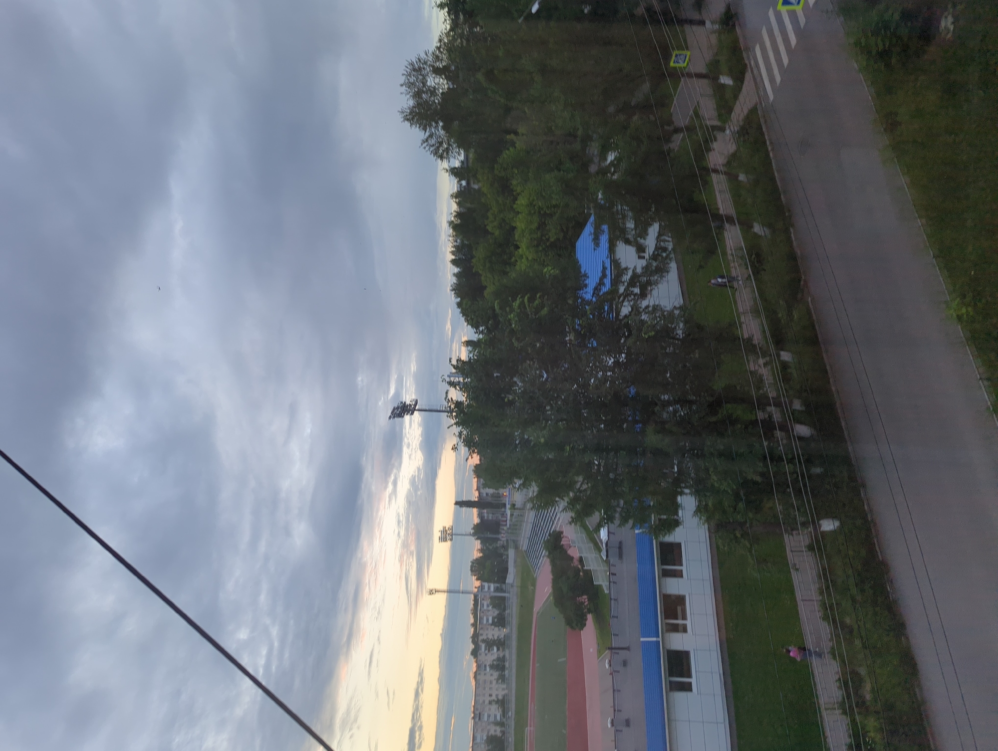 The image depicts an outdoor scene in Votkinsk, Russia, likely during the evening or early morning. In the foreground, a dark, diagonal line, possibly a wire or cable, crosses the frame. The background reveals a sports complex with a stadium featuring a running track and seating areas, illuminated by tall floodlights. Adjacent to the stadium is a low-rise building with a white facade and blue trim, featuring multiple windows. Further in the distance, a dense line of trees separates the sports facilities from a residential area characterized by multi-story apartment buildings. A paved road with a pedestrian crosswalk is visible in the middle ground, with a few individuals walking along a pathway. The sky is overcast with dramatic clouds, hinting at an approaching storm or sunset.