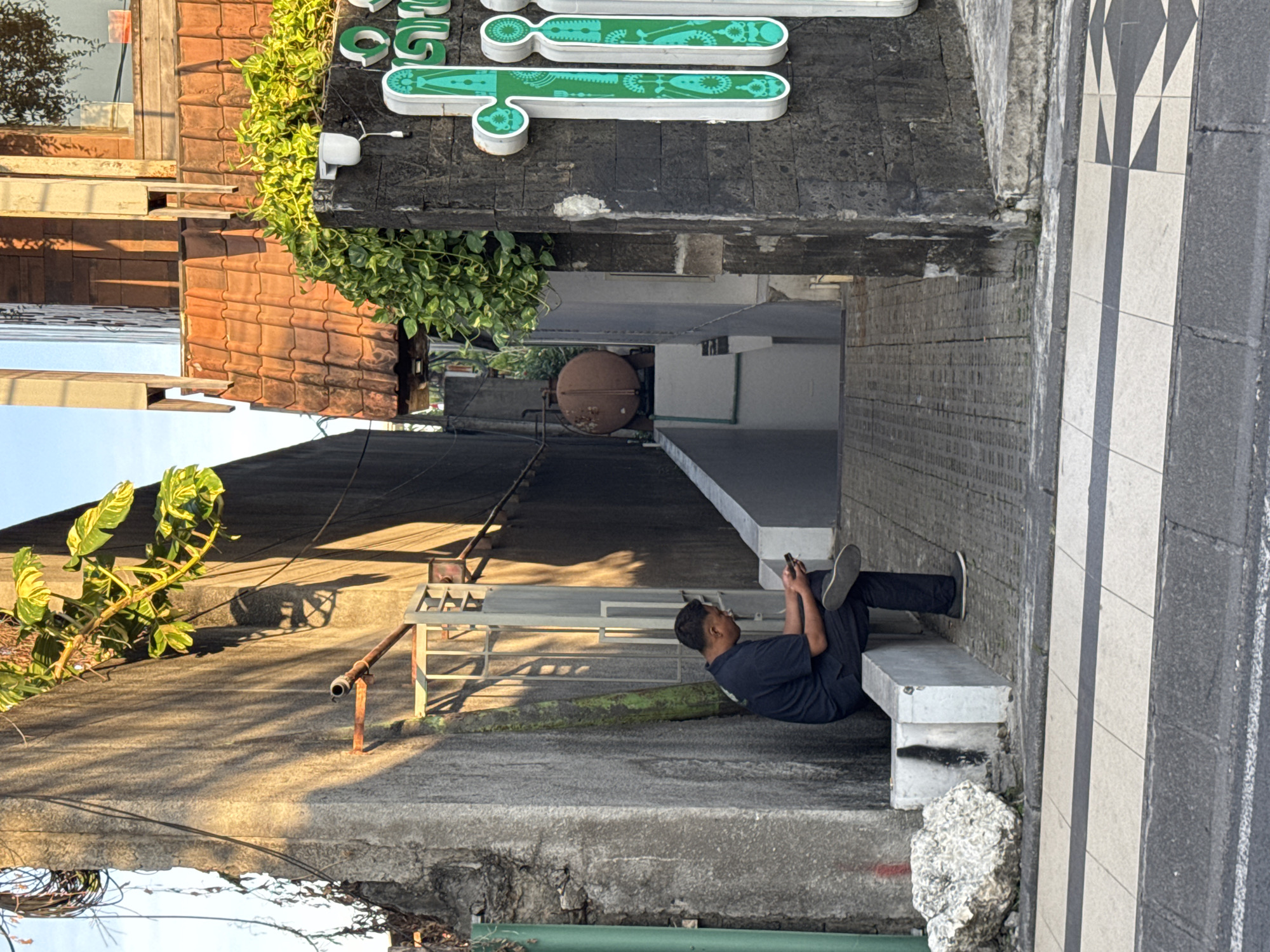 This urban scene, likely set in Kuta, Indonesia, captures a man relaxing on a concrete bench amidst a tapestry of buildings, greenery, and architectural details, illuminated by warm, late afternoon sunlight. The photograph presents a multi-layered view of a bustling, possibly alley-like, environment.
In the lower half of the frame, a man with dark hair, wearing a dark t-shirt, dark pants, and sneakers, is seated casually on a low concrete bench. He leans against a textured grey wall while engrossed in his smartphone, held in both hands. His pose suggests a moment of rest or waiting. To the right of the man, a wall or ground surface features a striking black and white geometric pattern of triangles and rectangles, hinting at a public or commercial area.
Above the man, various structures are visible. Terracotta-tiled roofs, characteristic of tropical architecture, are topped with lush green foliage that spills over the edges. Below these roofs, a darker building facade hosts a prominent green sign composed of stylized, elongated shapes outlined in white and adorned with intricate internal patterns, possibly spelling out a business name or brand. A small white security camera is mounted on this facade near the sign. Further into the background and at a slightly lower elevation, a large, rusty, spherical object, resembling a ventilation globe or water tank, sits on a flat roof.
The warm, golden light of the sun casts distinct shadows across the concrete surfaces, enhancing the textures of the walls and foliage. Various pipes, metal structures, and utility wires are discernible throughout the scene, adding to the urban character. The combination of specific architectural styles, abundant greenery, and the overall atmospheric lighting provides strong contextual clues for its Indonesian location.
