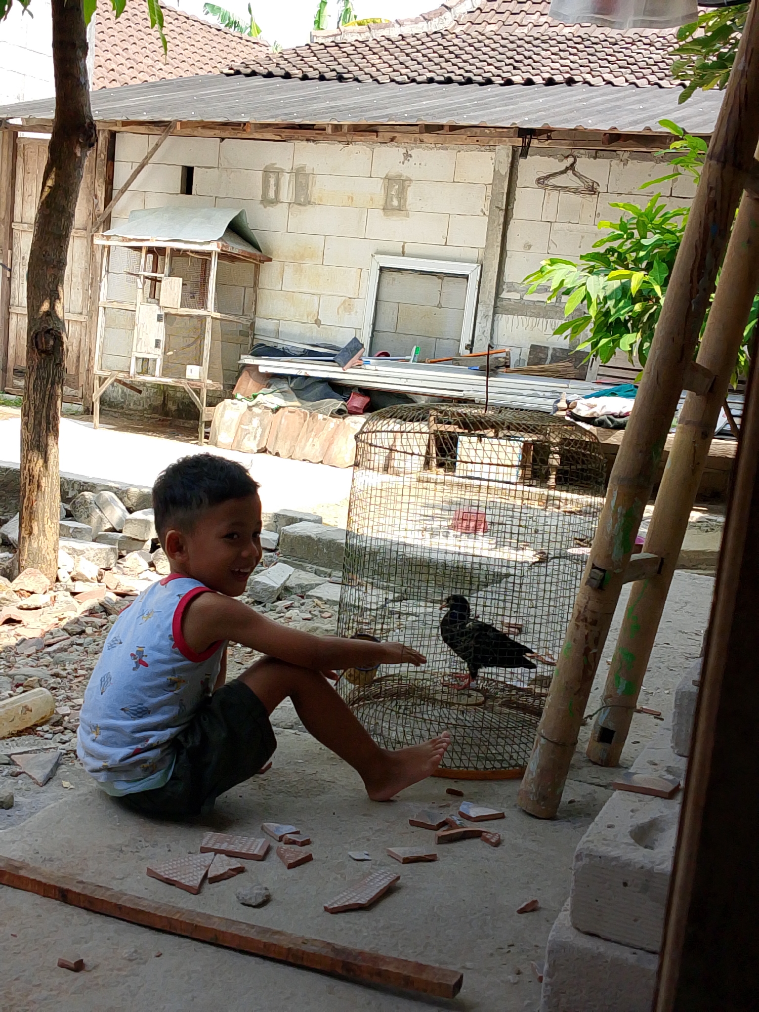 In Penyangkringan, Indonesia, a young boy with dark hair, wearing a light-colored tank top with a red trim and dark shorts, is sitting on a concrete ground littered with broken ceramic tiles and small rocks. He is smiling and looking to his right. To his right, a black bird with red feet is inside a circular wire birdcage. The birdcage is positioned next to a wooden structure made of bamboo poles. In the background, there is a building constructed from light-colored bricks, with a tiled roof. A smaller, wooden birdhouse-like structure is visible to the left of the main building. The environment appears to be an outdoor area, possibly a yard or alleyway, with overgrown vegetation in the upper right portion of the image.