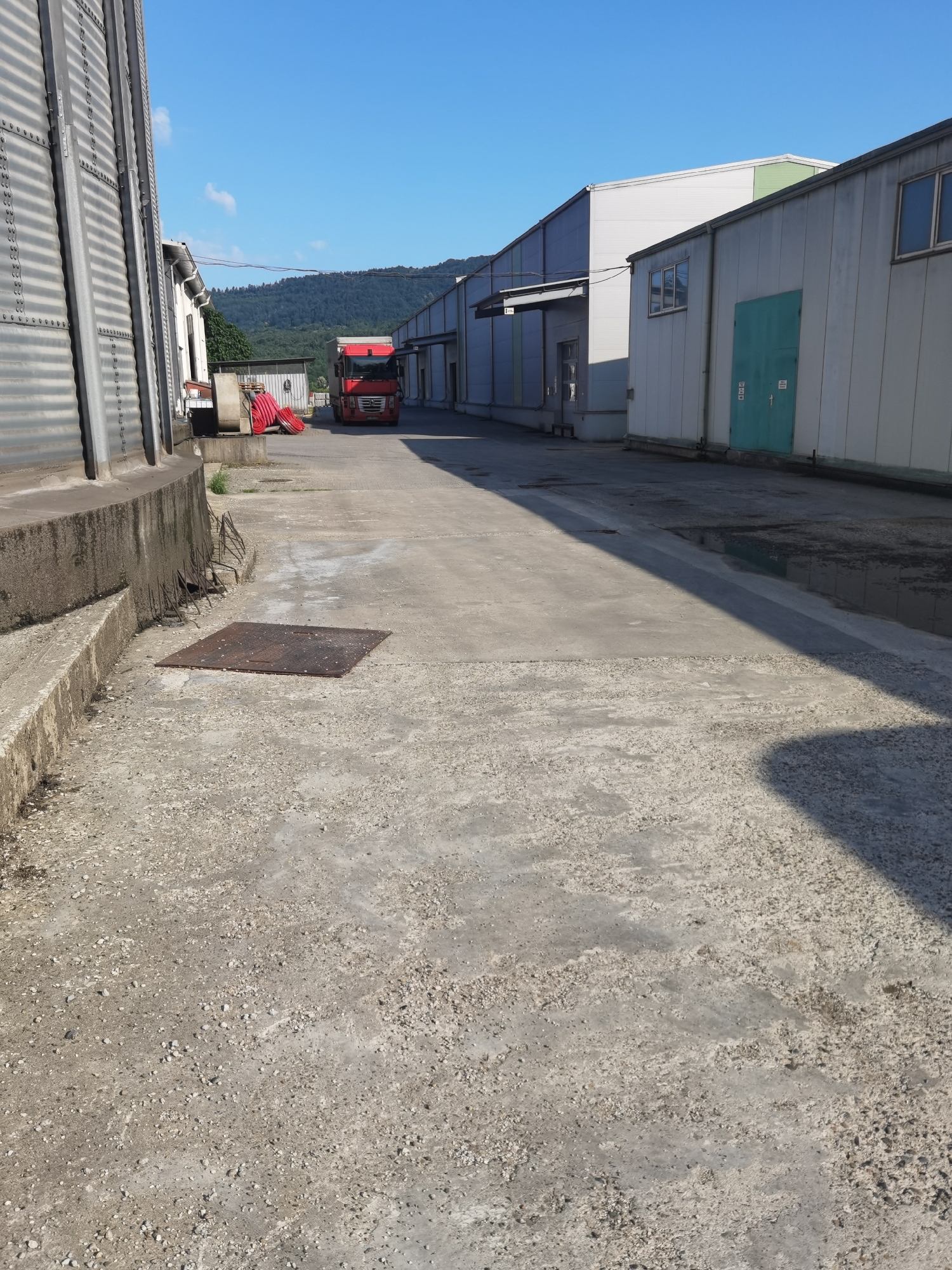 This is a medium-shot, eye-level, outdoor photograph taken on a sunny day in Lomets, Bulgaria. The scene depicts an industrial or agricultural area with several buildings and a red truck.
In the foreground, a rough, light-colored concrete ground surface is visible, with scattered small stones and patches of darker discoloration. To the left, a section of a tall, corrugated metal silo is prominently featured, with its concrete base running along the lower edge of the frame. In the middle ground, a square, rusty metal grate is embedded in the concrete.
Further into the scene, a red semi-trailer truck is parked. Behind the truck, there are piles of red coiled materials, possibly hoses or tarps, resting on a concrete platform.
The background consists of several industrial-style buildings with light-colored metal or panel exteriors. One building on the right has a prominent teal-colored door. Further back, a line of trees and hills can be seen against a clear blue sky. The overall impression is of a functional, possibly storage or processing facility.