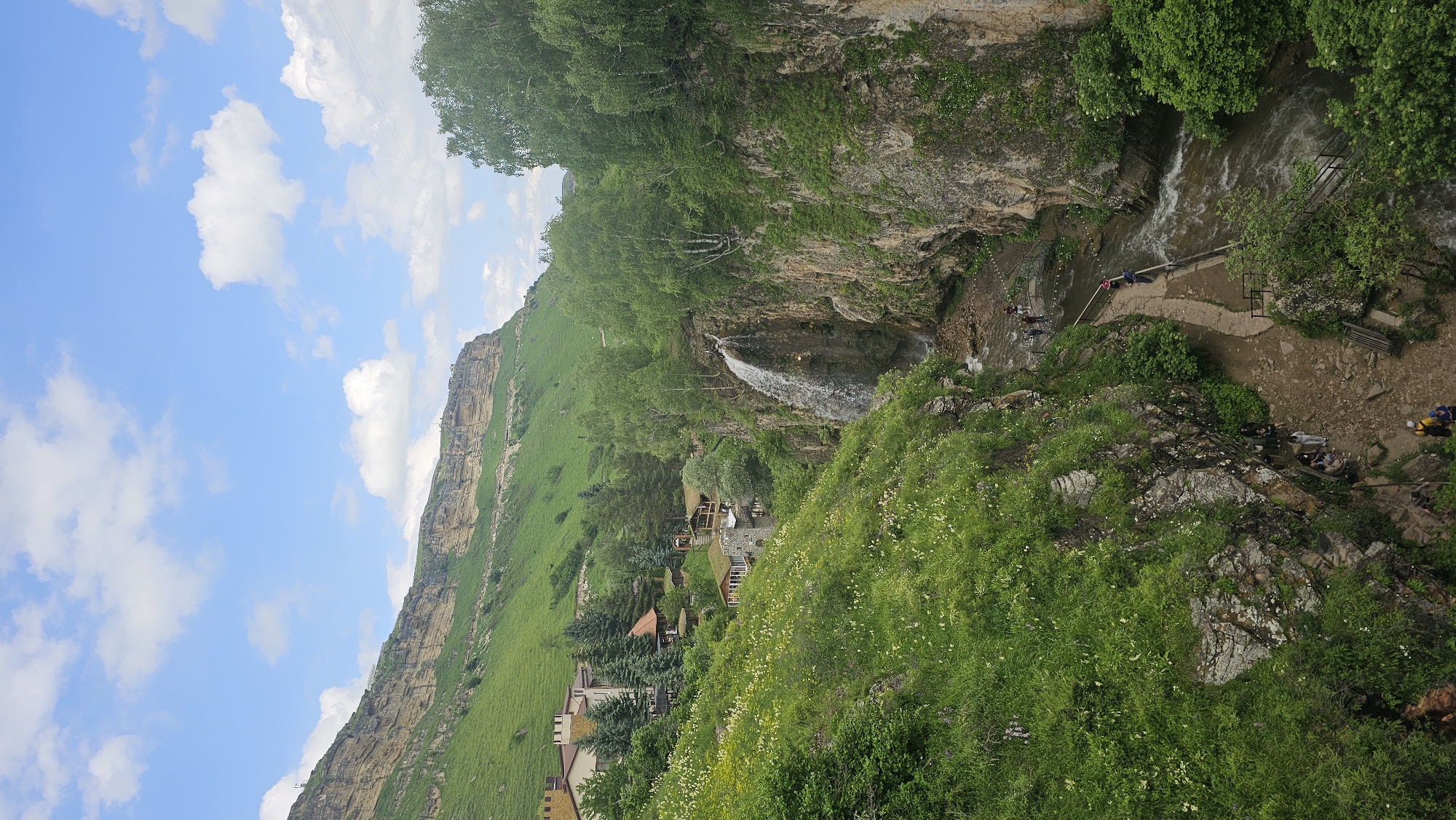 In a mountainous landscape under a partly cloudy blue sky, a waterfall cascades down a rocky gorge. On the left side of the gorge, a village is nestled amongst green hillsides. Buildings with distinctive red roofs and turret-like structures are visible. A winding path leads down into the gorge, with several people walking along it. The path is bordered by lush green vegetation and rocky outcrops. The waterfall feeds into a river that flows through the bottom of the gorge, with more people seen near the water's edge. The overall impression is a scenic natural location with a small settlement integrated into the landscape.