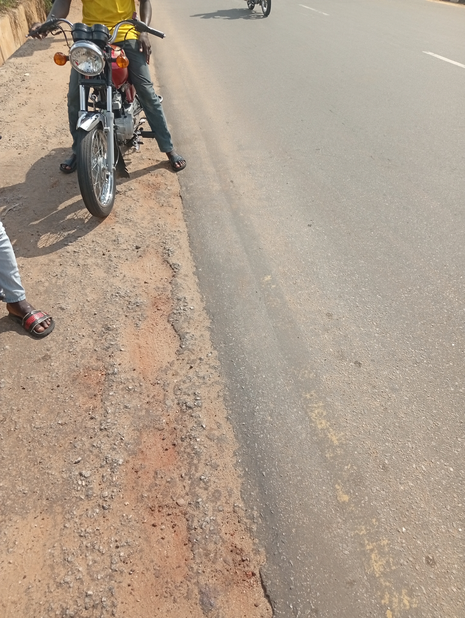 The image depicts a red motorcycle parked on the side of a road in Adankolo, Nigeria. The road is paved and bordered by a dirt shoulder. A person wearing a yellow shirt and jeans stands astride the motorcycle. A second person, partially visible, stands nearby. The environment appears to be daytime.