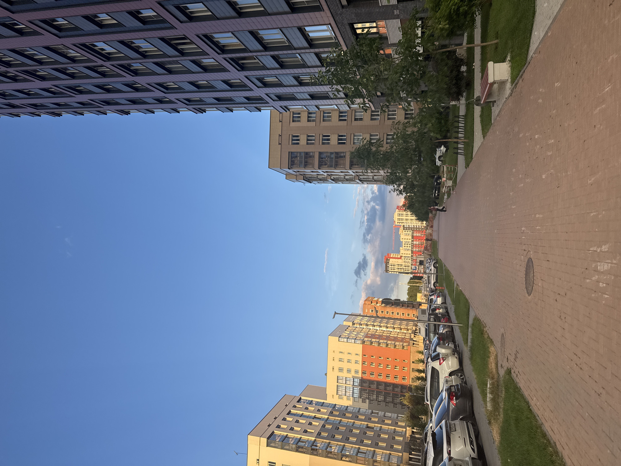 This is an outdoor, eye-level shot of a modern residential area in Koltsovo, Russia. The scene is dominated by a wide, reddish-brown brick paved walkway that extends into the distance, lined with green grass. To the left of the walkway is a row of parked cars, including white SUVs and sedans. Beyond the cars, several multi-story apartment buildings are visible, varying in color and architectural style, with a clear blue sky overhead. To the right of the walkway, a dark, modern apartment building stands tall, featuring rows of windows. Several trees with green foliage are planted between the walkway and the buildings, and a park bench is visible on the right side. In the distance, more apartment buildings and some construction cranes are present. A few indistinct figures are walking on the walkway. The lighting suggests it is late afternoon or early evening, with long shadows cast by the buildings.