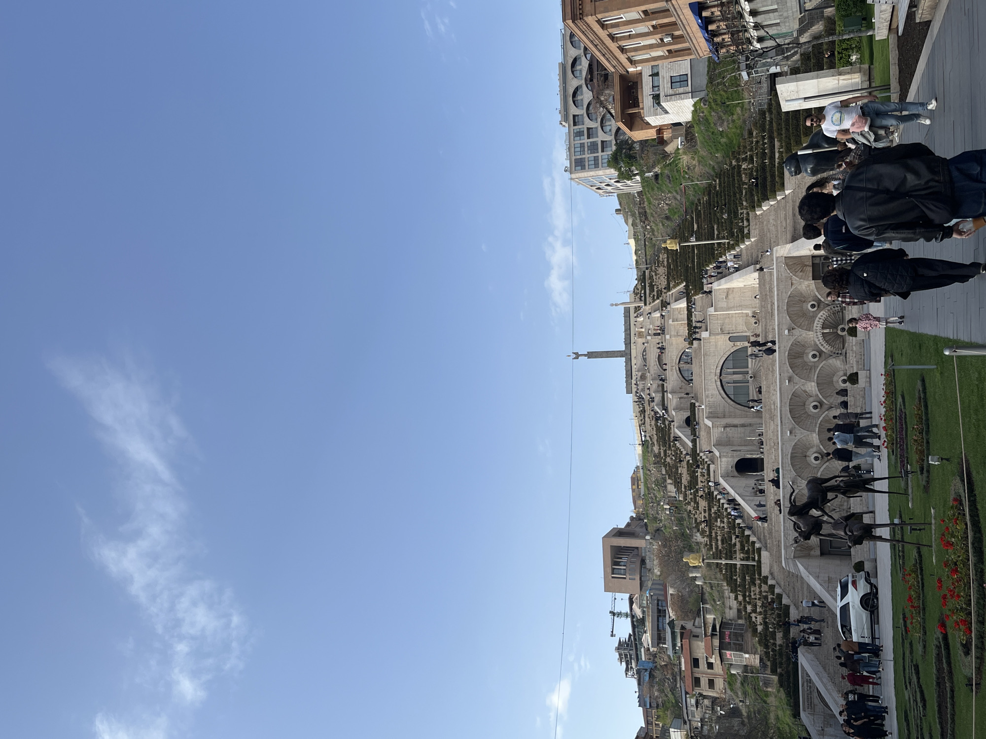 The image depicts the Yerevan Cascade in Armenia, viewed from an elevated position. The Cascade is a monumental stairway connecting the city center to the upper residential areas. The steps are adorned with fountains and sculptures. People are walking up and down the Cascade. Buildings are visible on either side. The sky is a clear blue with sparse clouds. A white car is parked near the bottom. There are also several statues and plant beds. A construction crane is visible further down the hill.