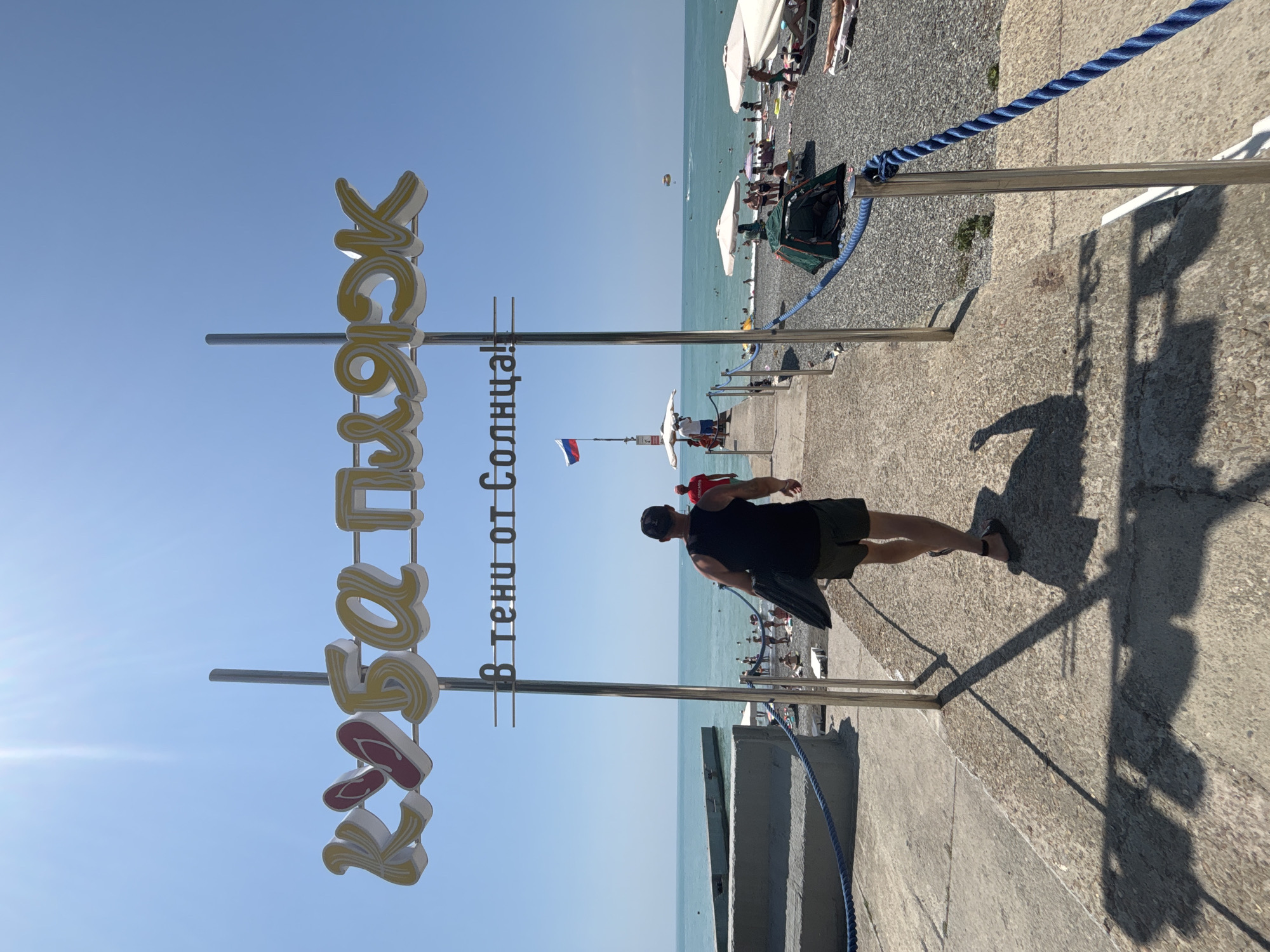 This is a photograph taken on a sunny day at a beach in Sochi, Russia. The image is framed by a concrete pathway leading towards the sea. Dominating the upper part of the frame is a large, yellow sign with stylized lettering, reading "КУБА ПЛЯЖ" (Kuban Beach). Below this, a smaller sign says "В тени от Солнца!" (In the shade from the Sun!). A Russian flag flies on a pole near the beach.
A man wearing a black tank top and khaki shorts is walking away from the camera on the pathway, carrying a dark object. Several people are visible on the beach, relaxing under umbrellas, sunbathing, and swimming in the turquoise sea. A small green tent is set up on the pebble beach. In the distance, a parasail is visible in the sky over the water. The overall scene depicts a typical summer day at a popular seaside resort.