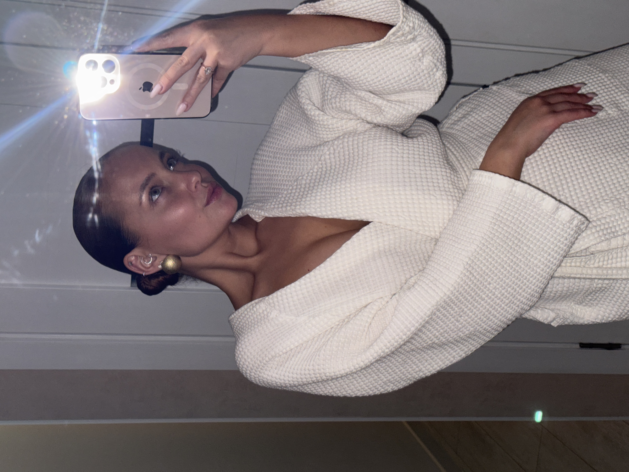 A woman with her hair in a bun is taking a selfie with her iPhone. She is wearing a white waffle-knit robe, and has a gold earring on one ear. She is wearing a ring on one finger. The background appears to be a room with light-colored walls. The location is not specified, and there is nothing in the image to indicate that it is Chaikovsky, Russia.