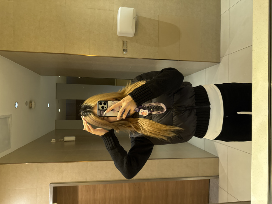 The image depicts a female individual's reflection in a mirror, situated in an indoor facility in Yuzhong District, China. The environment features light beige square wall tiles, white floor tiles, and a wooden-textured door panel visible on the left. Overhead, multiple circular recessed light fixtures are reflected. A white wall-mounted hand dryer is affixed to the right wall, beneath which a small rectangular sign is present. A white wall-mounted dispenser is located on the upper left wall. A grey countertop forms the base of the mirror.
The subject possesses long, layered hair, blonde from the mid-shaft to the ends with dark roots visible. Her facial features are obscured by a smartphone, held with both hands to capture the reflection. She is dressed in a black cropped puffer vest worn over a black knitted long-sleeved top, a white undershirt, and black trousers. A lanyard or decorative charm, featuring an orange and white emblem, is suspended from the smartphone. The individual's right hand is elevated, with fingers positioned near the crown of her head, while her left hand supports the phone during the self-portrait capture.