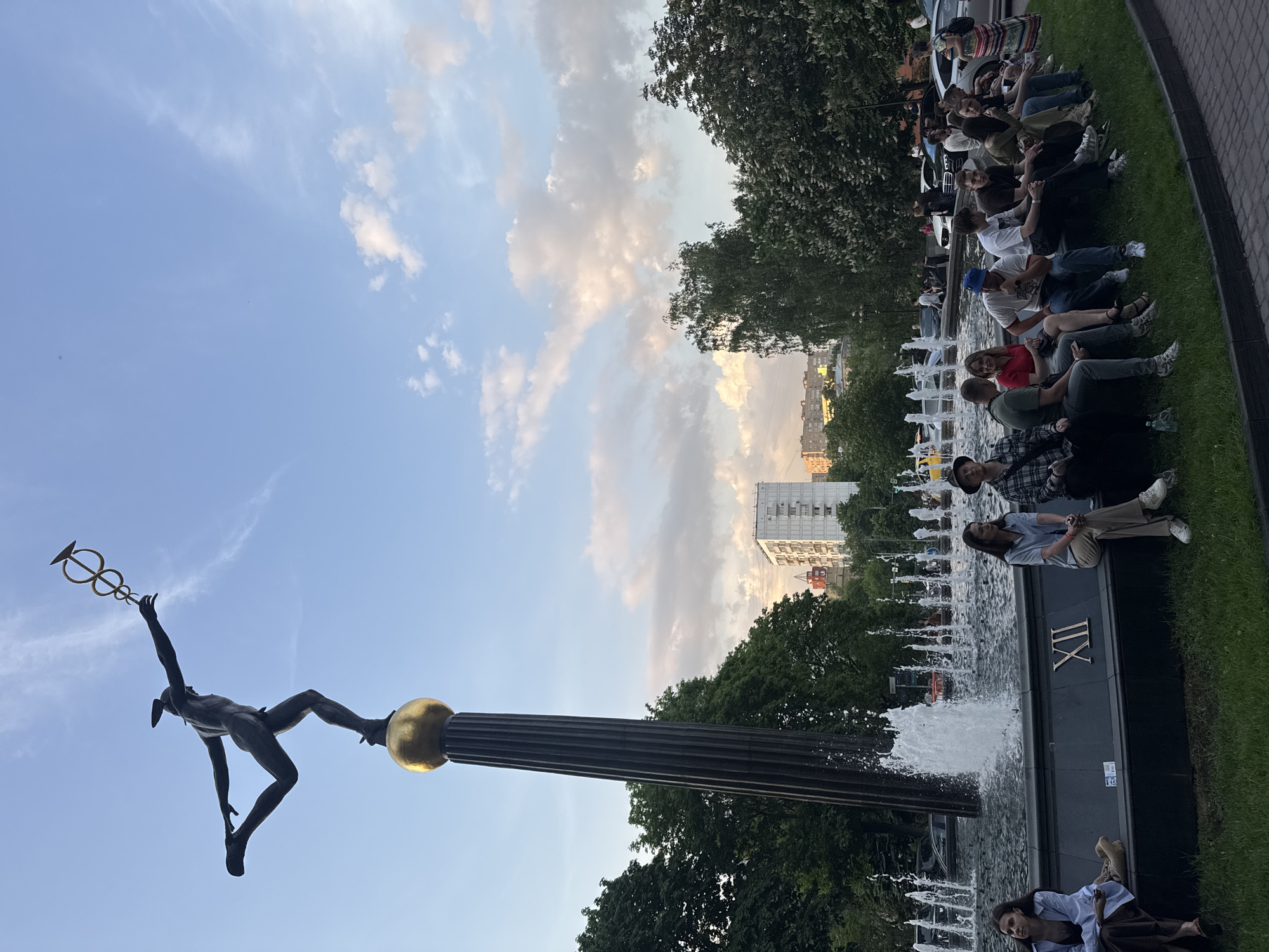 A wide-angle, upward-tilted shot shows a public gathering at the 'Four Seasons' fountain in Manezhnaya Square, Moscow. A bronze sculpture of a naked figure, holding a caduceus, is mounted atop a fluted column capped with a golden sphere. This is positioned to the left of the fountain, which features Roman numerals on its base.
To the right of the fountain, a group of people is sitting on the grass. They appear to be observing the fountain or each other.
Behind the fountain and people, lush green trees fill the midground. In the background, multi-story buildings are visible under a partially cloudy sky, hinting at late afternoon or early evening light.