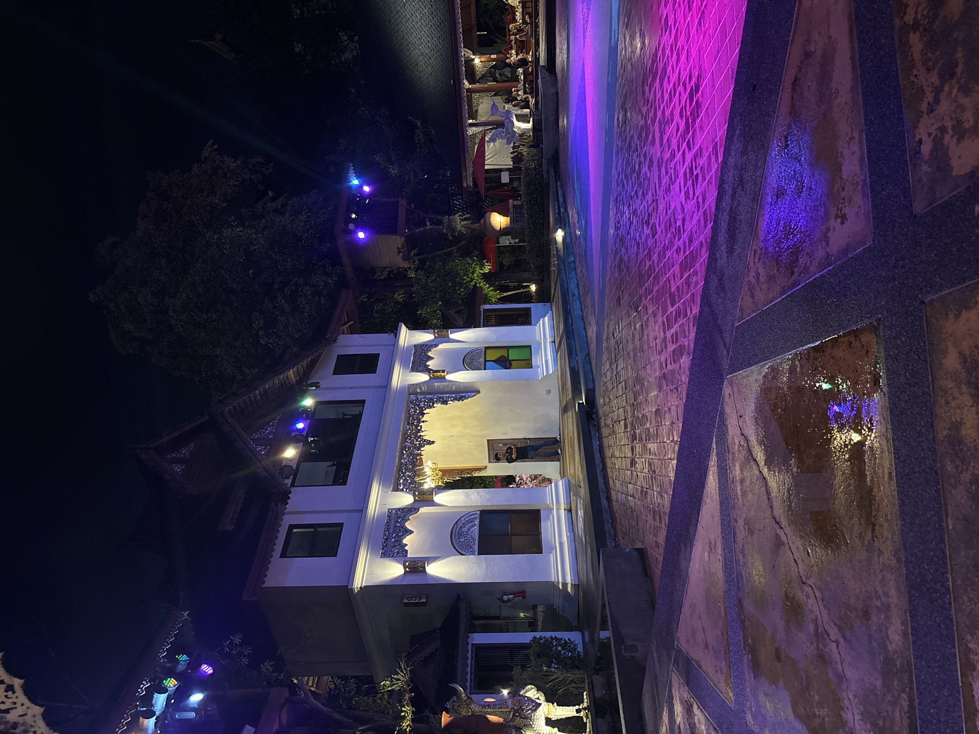 An outdoor night scene depicts a paved courtyard with a building and trees. The building, predominantly white with traditional Thai architectural elements, features ornate carvings above its entrance and windows. A man in a dark shirt and pants stands near the building's entrance. Lights, some colored purple and blue, illuminate the scene from above and the building's facade. The courtyard floor is paved with bricks and some sections appear wet, reflecting the colored lights. In the background, there are more structures, red umbrellas, and a seated group of people at tables. The overall atmosphere is festive and illuminated, suggesting an event or entertainment venue in Nong Pa Khrang, Thailand.