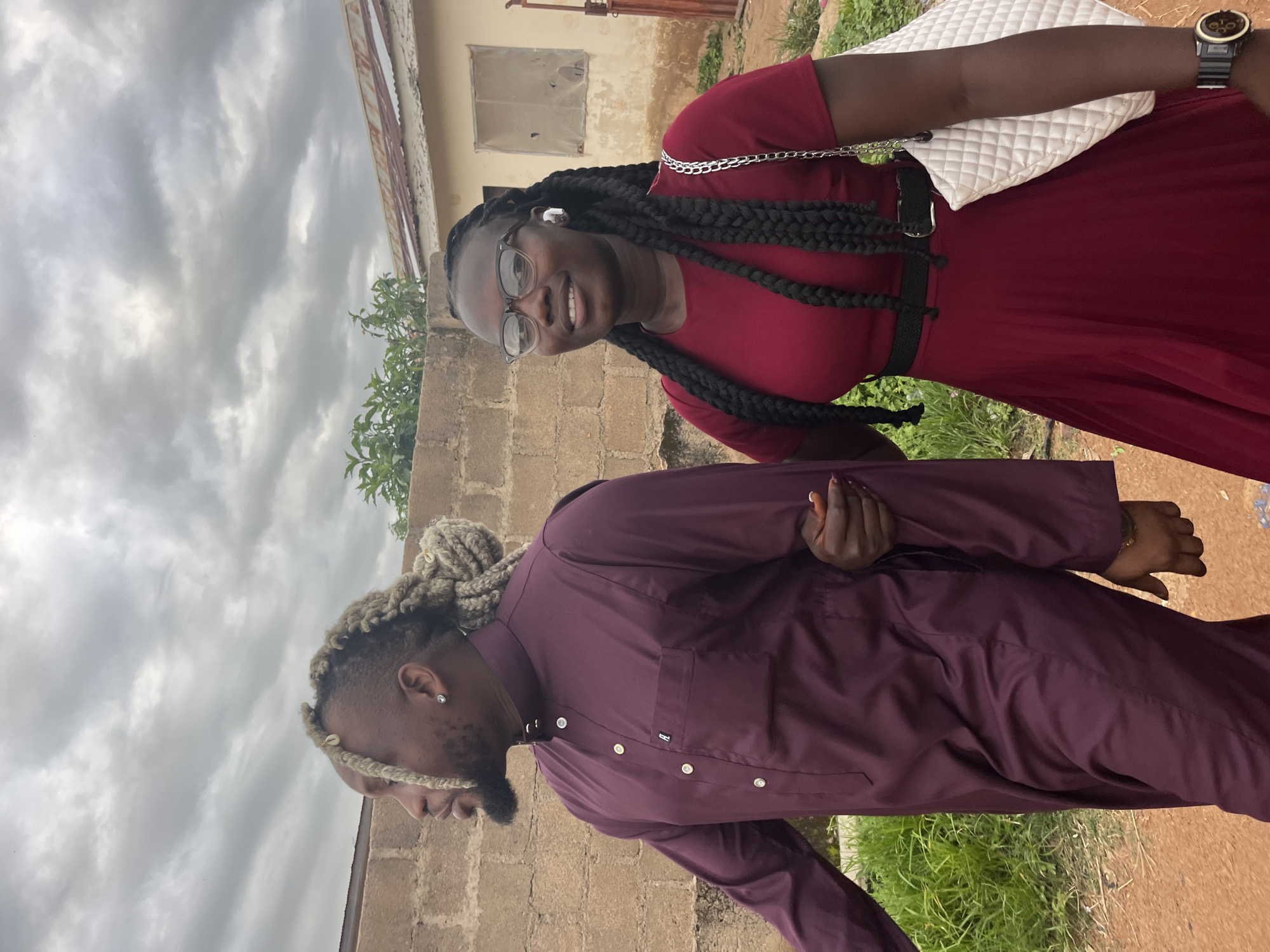 A man and a woman are walking side-by-side in front of a brick wall and a building in Jos, Nigeria. The man, on the left, has braided blonde hair and is wearing a burgundy traditional outfit. The woman, on the right, has long black braids, glasses, and is wearing a burgundy dress. She is holding a white quilted handbag. The sky is cloudy, and there is vegetation and dirt ground visible.