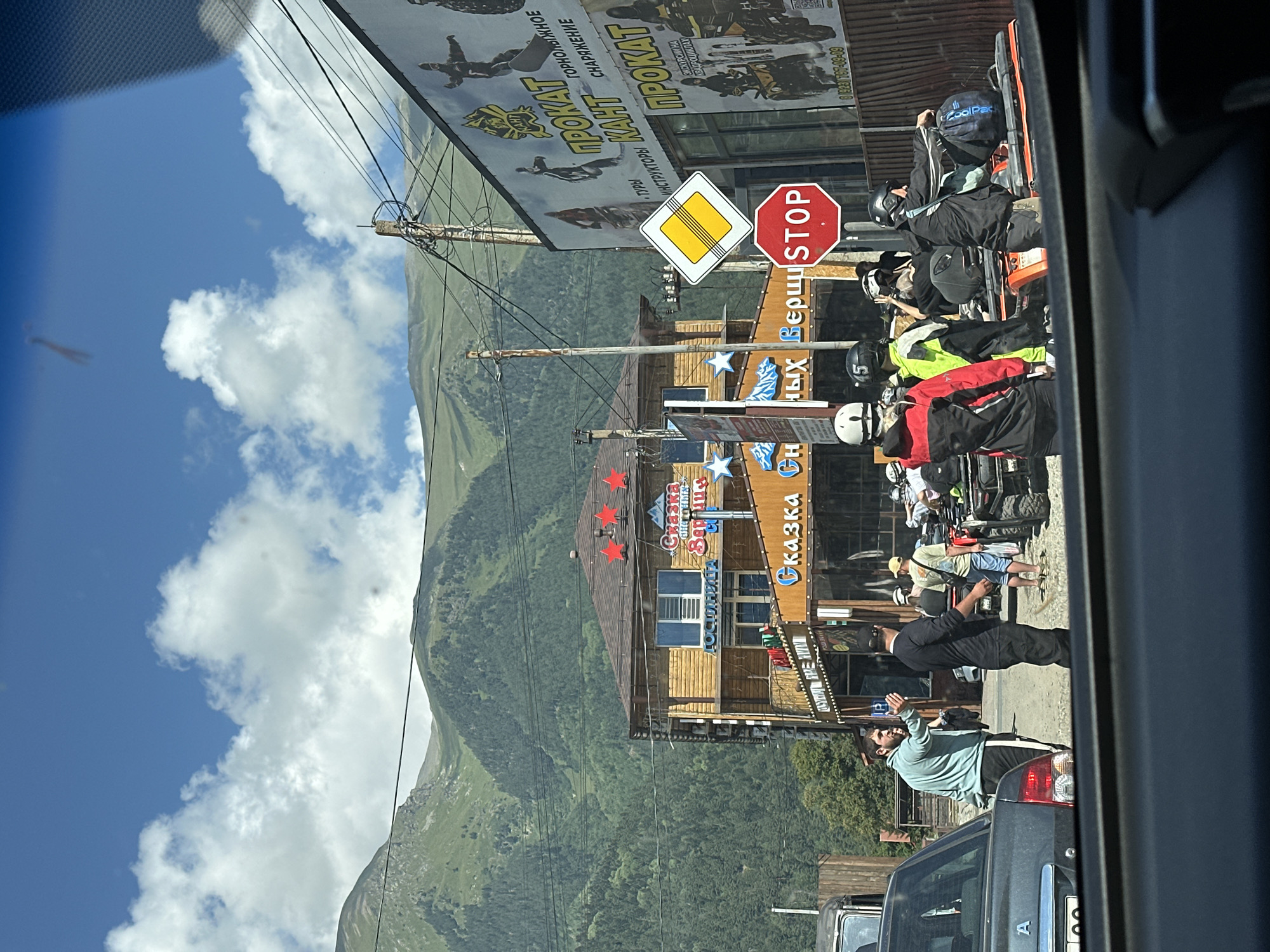 The image captures a street scene in Arkhyz, Russia, likely a tourist area given the surrounding mountains and activity. A multi-story wooden building with red stars and Cyrillic signage, possibly a hotel or restaurant, stands prominently. In front of it, several people are gathered, some wearing helmets and geared up for activities like ATV riding, which is suggested by several ATVs parked nearby. One man in a light blue jacket is gesturing with his hand, seemingly interacting with another man in black. Various advertisements for "Prokat" (rental) are visible on a building to the right, displaying images of people on ATVs and snowboards. Road signs, including a "STOP" sign and a yellow diamond-shaped sign, are also present. The background is dominated by lush green mountains under a partly cloudy blue sky. The photograph appears to be taken from inside a vehicle, looking out through the windshield.