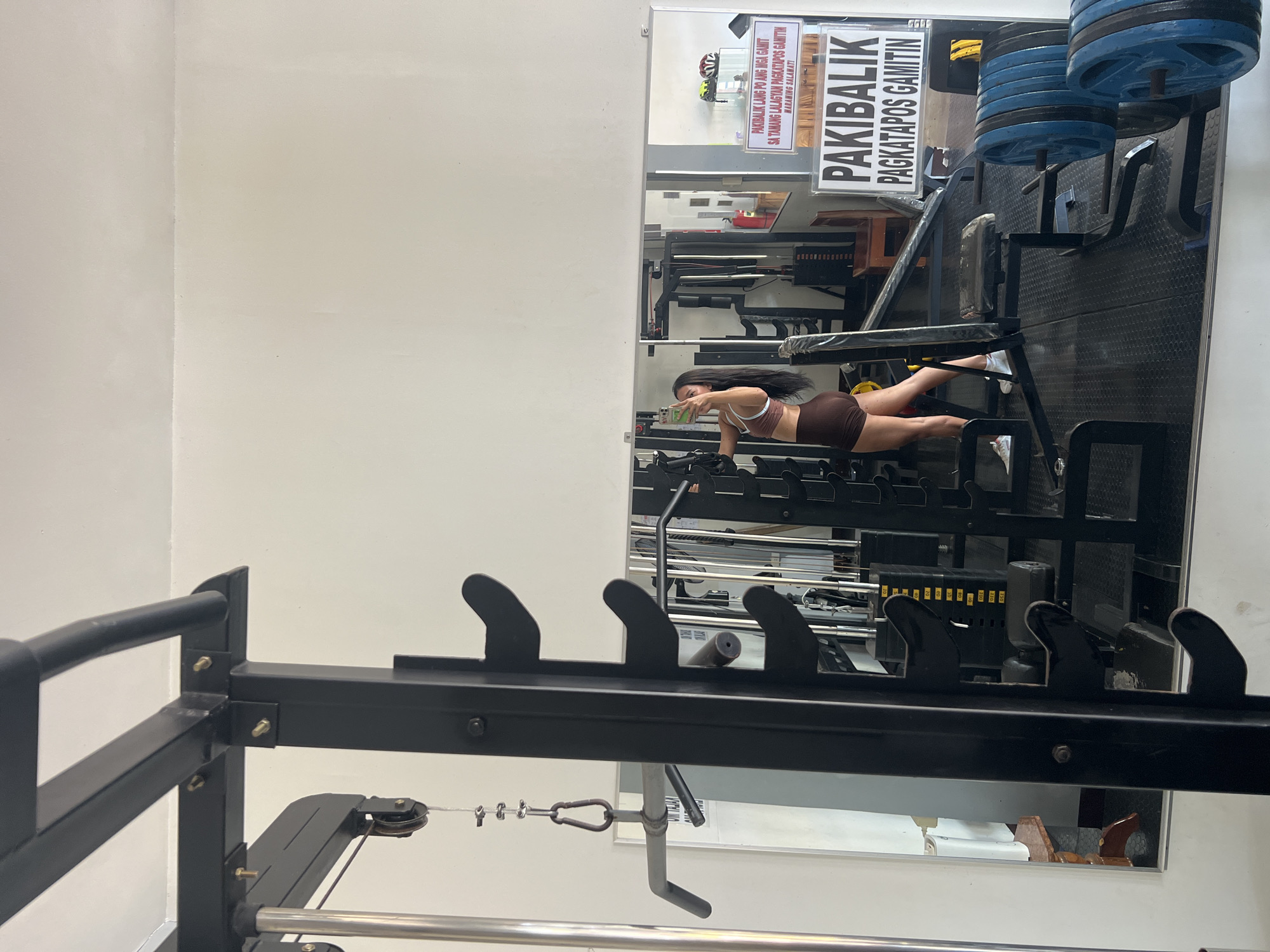 A woman is taking a selfie in a gym. She is wearing a brown sports bra and shorts. The gym is equipped with various exercise machines and weights. There are signs on the wall in the background with text that appears to be in Filipino. The location is specified as Cuyo, Philippines.