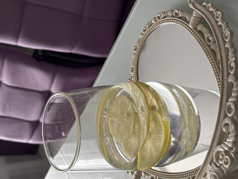 A clear, cylindrical glass containing water and multiple yellow lemon slices is positioned on a small, oval-shaped mirror tray with an ornate, light-colored frame. The glass and tray rest on a light-colored, reflective table surface. In the blurred background, two purple, tufted chairs are visible. The scene is indoors. No individuals are present. The location is Unknown, Unknown.