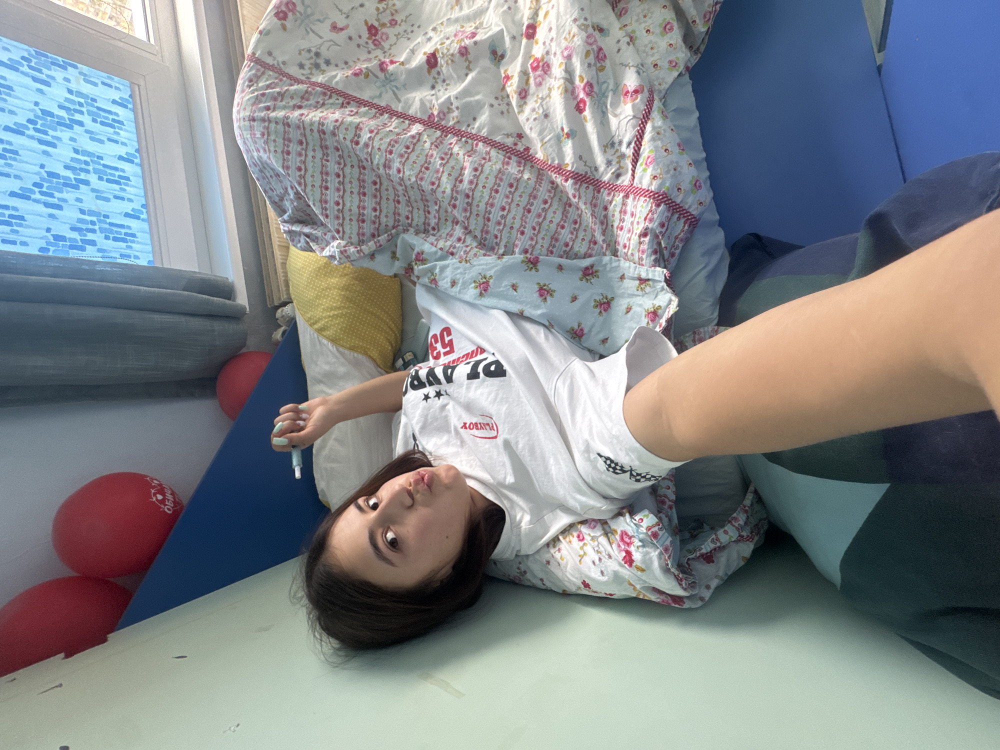 A young female is depicted lying on her back, looking directly at the camera with a playful pout. Her head is positioned towards the left side of the frame, with her body extending to the right. She rests on a light green surface, possibly a floor or mat.
Her left hand, adorned with light blue nail polish, holds a small white object, which appears to be a pen or a similar small tool. She is wearing a white t-shirt with visible text: "PLAYBOY" is printed upside down, and the number "53" is seen sideways, along with two stars and a black and white checkered pattern on the sleeve.
Behind and around her, bedding with a floral pattern of pink roses on white and light blue fabrics is visible, featuring sections with pink stripes. A yellow polka-dot pillow is also part of the bedding. To her right, a blue padded surface or wall extends upwards, adjacent to some darker blue and green bedding.
In the background, near the left side of her head, several red balloons are clustered against a blue surface. Partially obscured Cyrillic text is visible on one of the balloons. Further to the left, a white-framed window is visible, with a blue patterned blind or curtain seen through the glass, indicating bright daylight.
The setting appears to be an indoor space, likely a bedroom, conveying a casual and relaxed atmosphere. The provided context indicates the location is Sofia, Bulgaria.