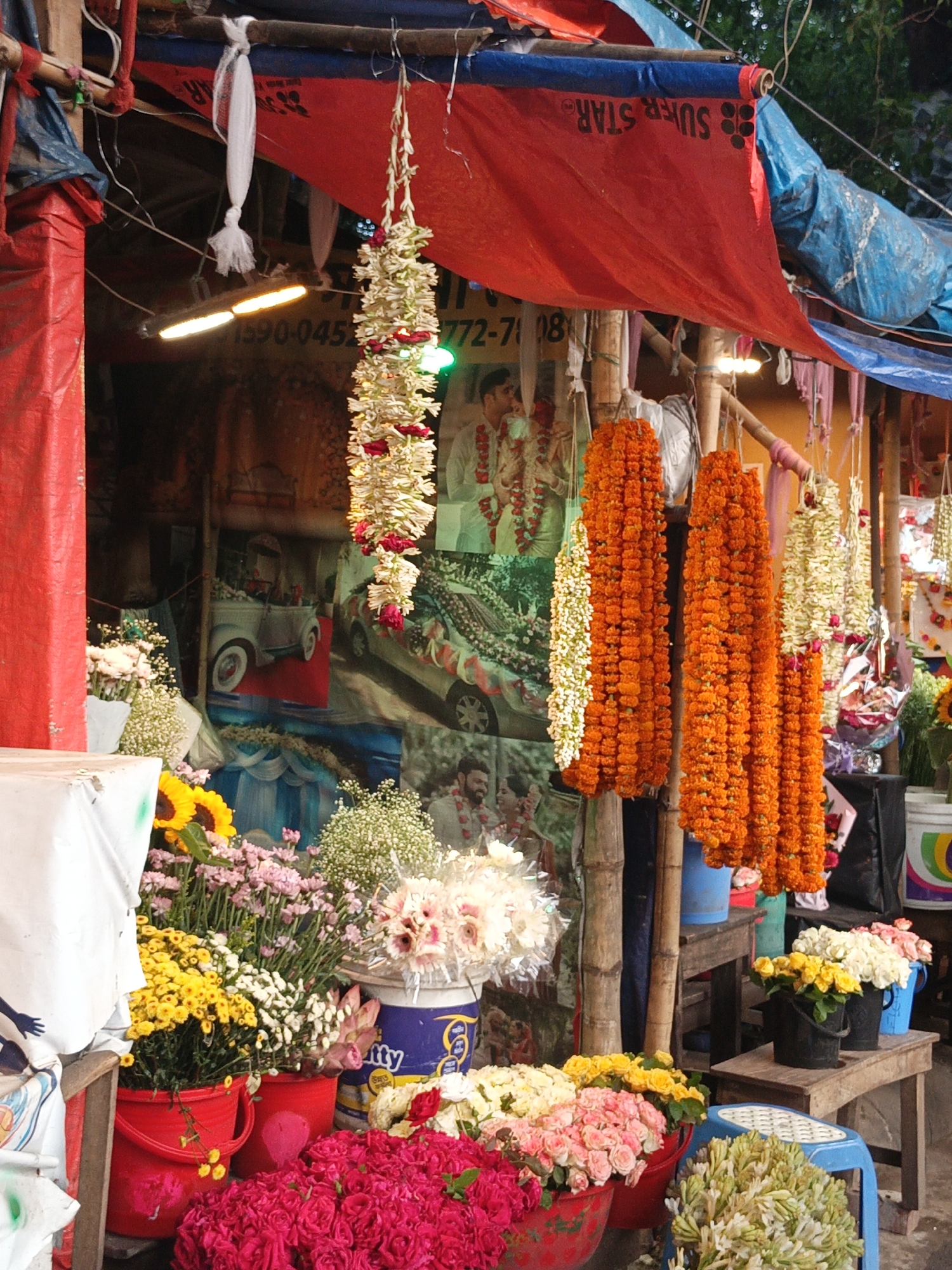 The image depicts a flower stall in Dhaka, Bangladesh. The stall is covered with a red and blue tarp bearing the word "Super Star." Garlands of orange and white flowers hang from the stall's roof. Various flowers, including roses, sunflowers, and chrysanthemums, are displayed in buckets and pots at the stall's front. Behind the flowers, posters depicting wedding couples and decorated cars are visible. The setting suggests a vibrant market scene.