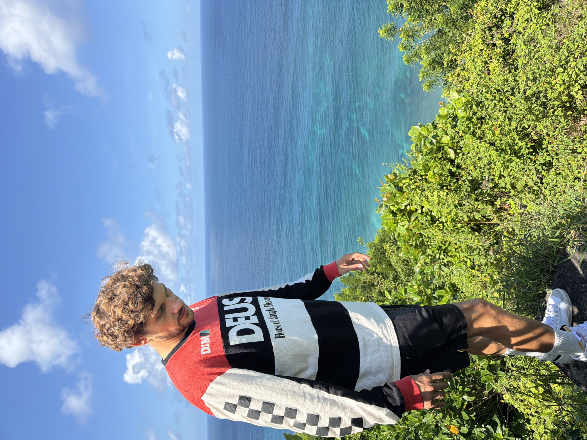 A full-body shot shows a man standing on the edge of a cliff overlooking a body of water. The man has curly hair and is wearing a long-sleeved shirt with the words "DEUS House of Simple Pleasures" written on it, black shorts, white socks, and white sneakers. He stands on top of lush green vegetation. Behind him is a body of blue water and a clear blue sky with some white clouds. The scene appears to be captured during daylight hours. It is in the city of Kuta Selatan, Indonesia.