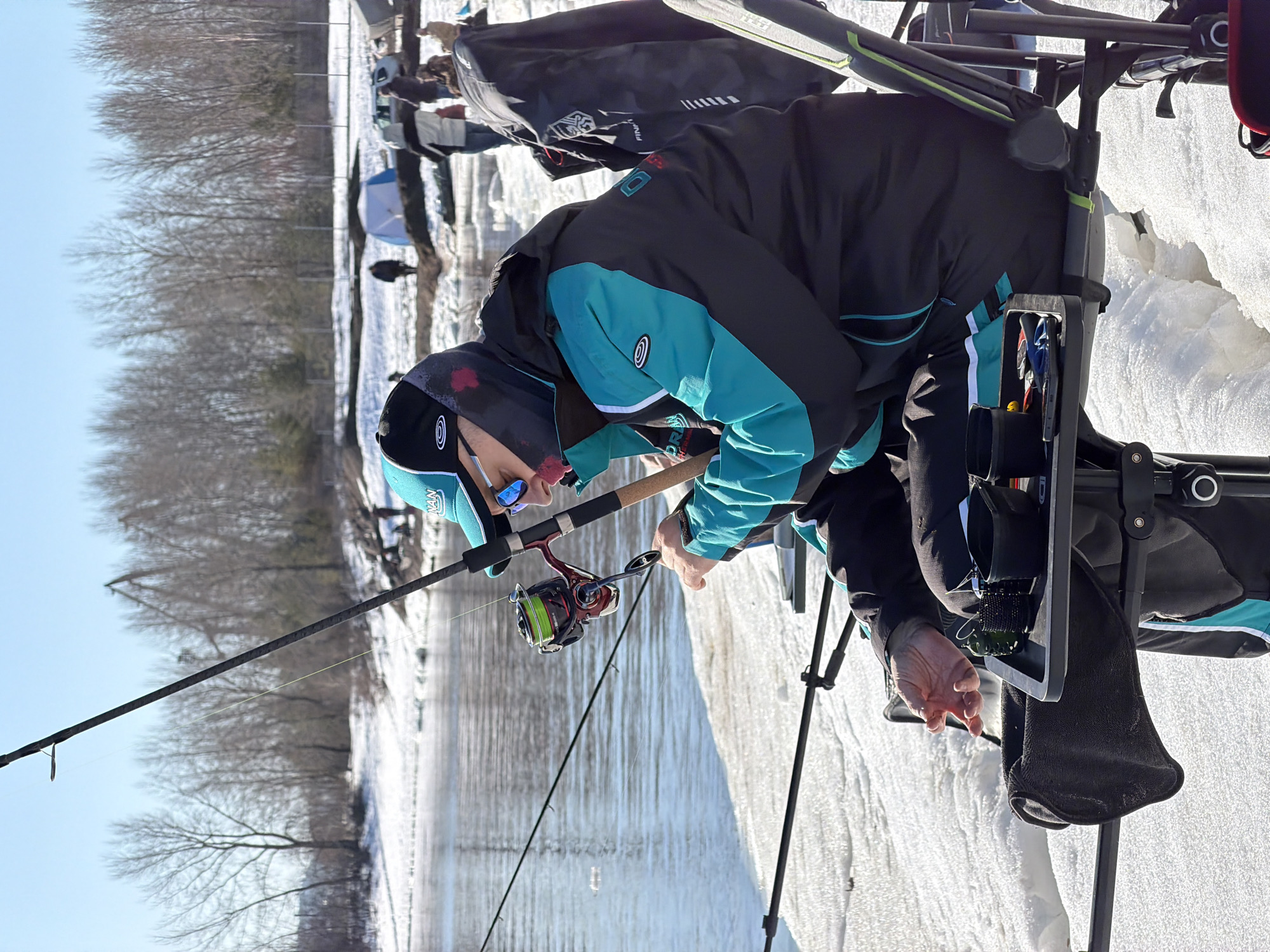 A man is fishing on the ice in Chaikovsky, Russia. The man is wearing a black and teal jacket, a black hat with a teal brim, and sunglasses. He is sitting in a black chair with a tray attached to it. He is holding a fishing rod with a red and black reel. The fishing line is in the water. There is snow on the ground and trees in the background. Other people are fishing nearby, with tents and equipment visible. The sky is clear and blue.