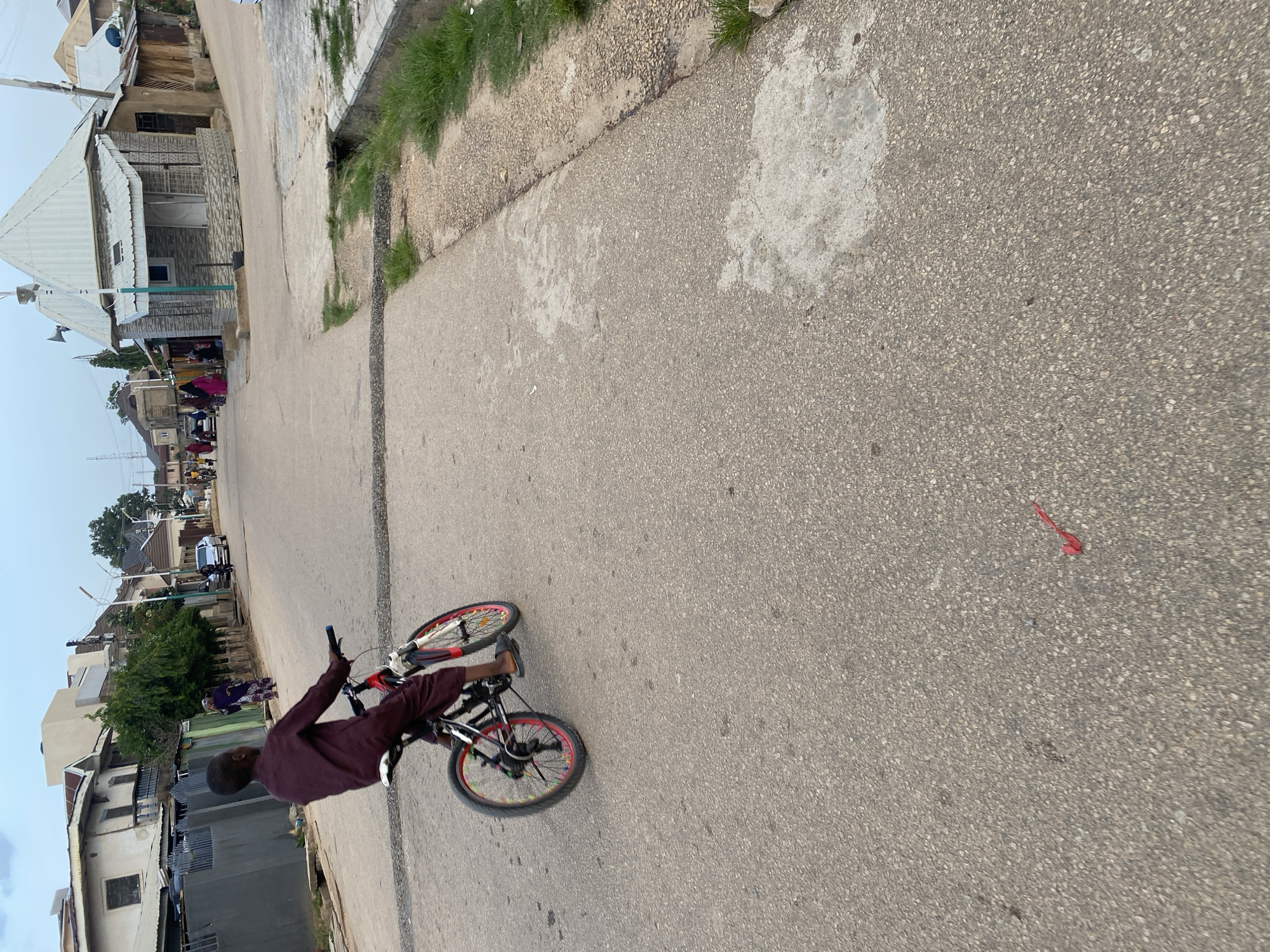 The image shows a young boy riding a bicycle in a street in Jos, Nigeria. The boy is wearing a maroon long-sleeved shirt and dark shorts. He is on a paved road with some debris and grass growing along the edge. Buildings, some appearing to be residences or shops, line the street on the left side. In the background, other people and a vehicle are visible, suggesting a community setting. A red ribbon or piece of fabric is lying on the road in the foreground. The overall lighting suggests it might be late afternoon or early evening, and the sky is clear. The scene appears to be a typical day in a Nigerian town. No visible text is present in the image.