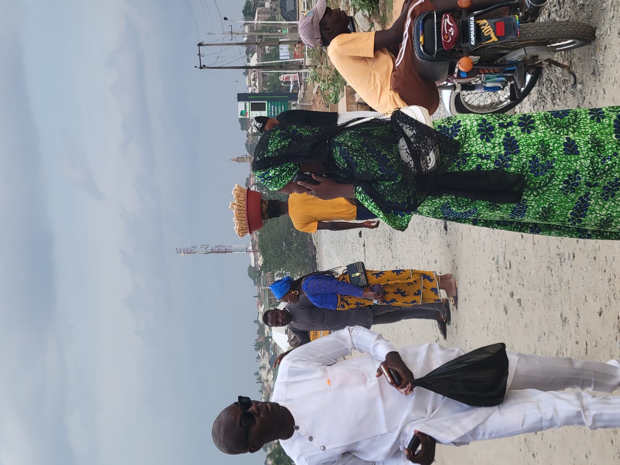 In Kwamba, Nigeria, several individuals are present. One person wears a white outfit with sunglasses. Another individual is atop a motorcycle. A woman in a green patterned dress is also visible, and another woman has a blue headscarf and a patterned yellow skirt. A person has a load atop their head. The setting appears to be outdoors, possibly along a roadside. There are buildings and power lines in the background.