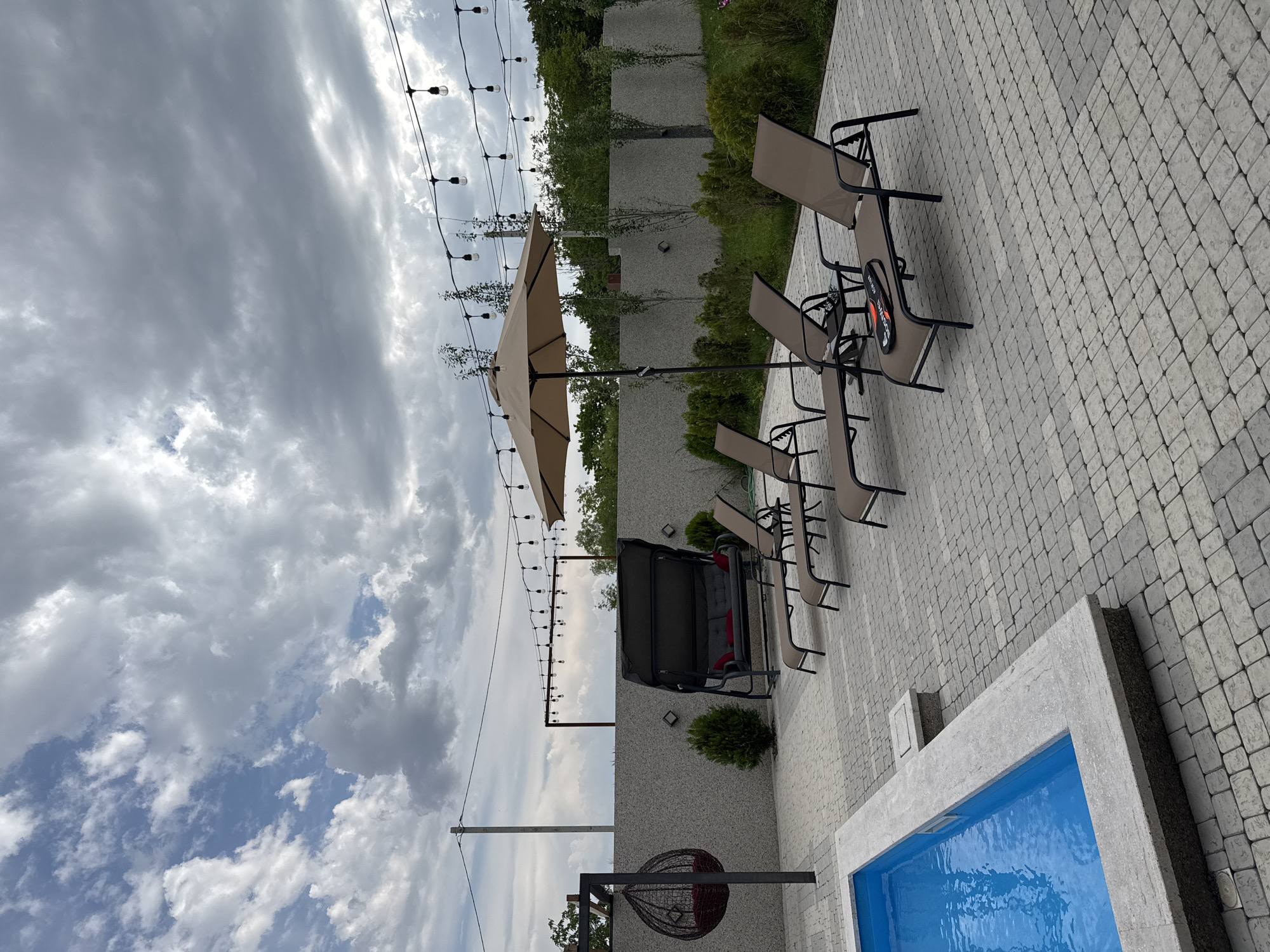 The image depicts an outdoor patio area adjacent to a swimming pool, likely located in Mughni, Armenia. The patio is paved with light gray interlocking stones. Several lounge chairs made of dark brown fabric and metal frames are arranged on the patio, with one chair in the foreground displaying a pair of rackets. A large, light brown patio umbrella stands open near the lounge chairs. In the background, a dark gray swing set with red cushions is visible against a textured gray wall. To the left, a dark circular hanging chair with a red cushion is suspended from a metal frame. A portion of a bright blue swimming pool with a light gray stone border is visible in the lower left corner of the frame. String lights are suspended across the upper portion of the image, and a dense green hedge borders the far side of the patio. The sky is overcast with dramatic, gray clouds.