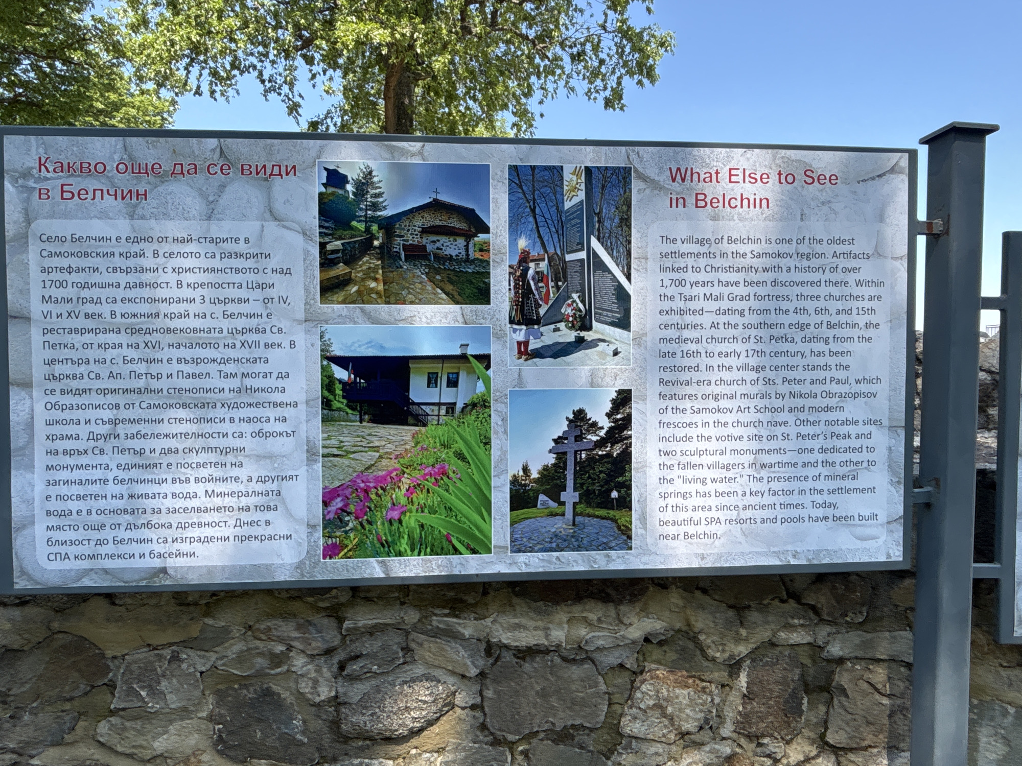 An outdoor information board is prominently displayed, detailing points of interest in Belchin, Bulgaria. The board features two textual panels, one in Bulgarian on the left and an English translation on the right, both titled "What Else to See in Belchin." Separating these panels is a 2x2 grid of four photographic images.
The images depict:
1. A stone church with a reddish-brown tiled roof, nestled on a sloped terrain with a paved pathway leading to its entrance, partially obscured by tree branches.
2. A person in traditional Bulgarian folk costume, including a tall feathered headdress, standing facing an angular, dark-colored monument with a golden emblem at its apex and a floral wreath placed at its base.
3. A close-up view of a house's veranda and a garden bed bursting with pink and purple flowers, with a stone path visible in the foreground.
4. A large stone cross monument erected on a paved base, surrounded by green foliage and pine trees under a clear blue sky.
The English text panel describes Belchin as one of the oldest settlements in the Samokov region, noting the discovery of over 1700-year-old Christian artifacts. It highlights three exposed churches within the Tsari Mali Grad fortress (dating from the 4th, 6th, and 15th centuries), the restored medieval church of St. Petka (late 16th to early 17th century), and the Revival-era church of Sts. Peter and Paul, featuring original murals by Nikola Obrazopisov. Other attractions include a votive site on St. Peter's Peak and two sculptural monuments—one dedicated to villagers fallen in wartime and another to "living water." The text concludes by mentioning the historical significance of mineral springs and the presence of modern SPA resorts and pools near Belchin.
The sign itself is mounted on dark grey or black metal posts. Behind the board, the upper branches of a large deciduous tree with lush green leaves are visible against a clear blue sky. The base of the sign is positioned in front of a rustic stone wall constructed from irregularly shaped, light-colored stones.