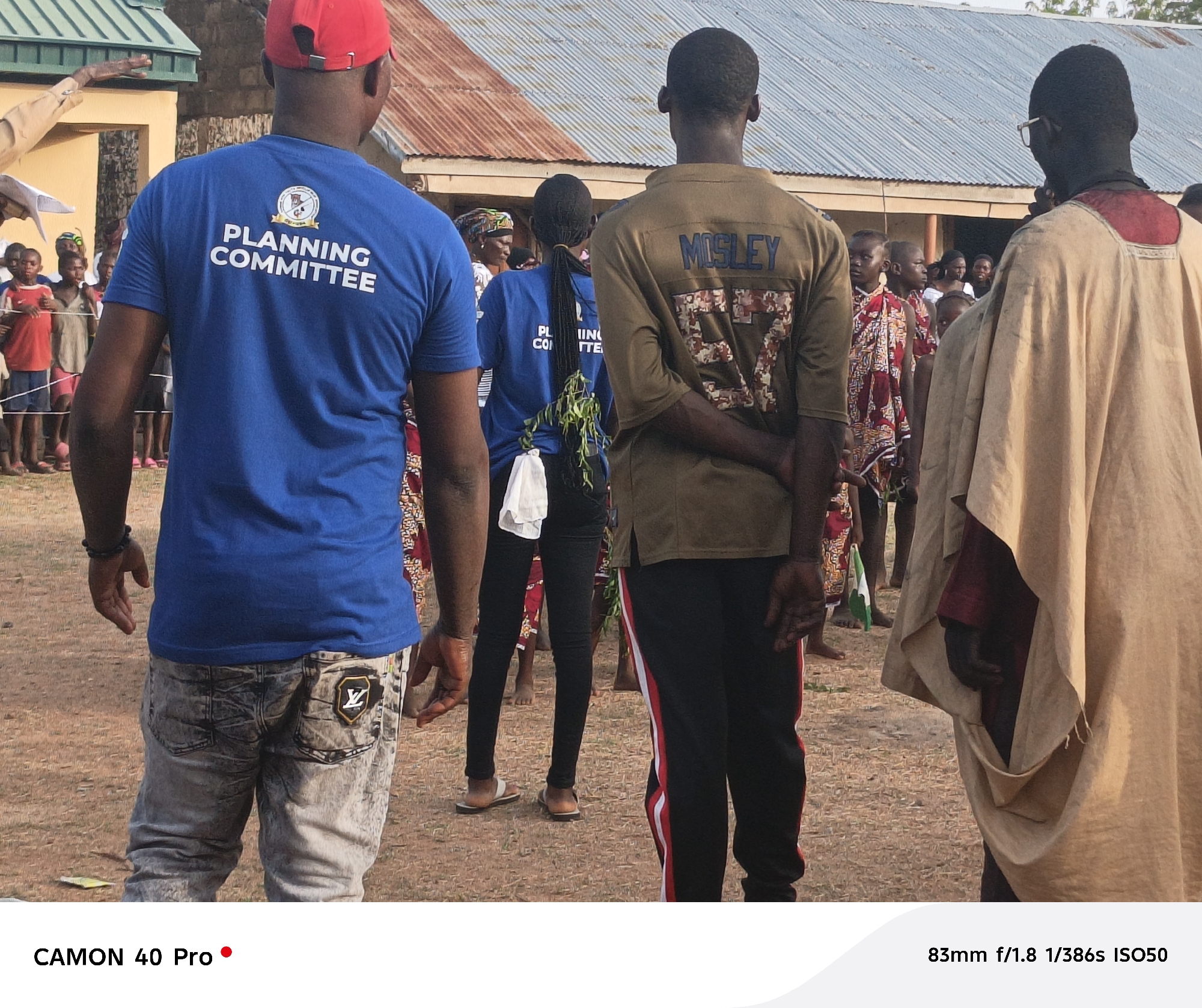 Here is a detailed description of the image:
The image shows a group of people standing in an outdoor area, likely in Langtang, Nigeria. In the foreground, four people are facing away from the camera. The first person on the left is wearing a blue t-shirt with "PLANNING COMMITTEE" printed on the back, along with light-colored jeans that have a logo on the back pocket. The second person from the left is wearing a blue shirt with what appears to be "PLANNING COMMITTEE" partially visible, paired with black leggings and sandals. They have long hair in a ponytail and are holding green foliage. The third individual is wearing an olive-green shirt with "MOSLEY" and the number "57" printed on the back, along with black pants that have a red and white stripe. On the rightmost side, a person is draped in a light beige garment and is wearing glasses.
Behind these four individuals, a crowd of people, including children and adults, is gathered, and there is a low building with a corrugated metal roof in the background. The ground is covered in dirt. The image also features camera information at the bottom, indicating the camera model is a "CAMON 40 Pro" and providing the technical details of the shot (83mm f/1.8 1/386s ISO50).