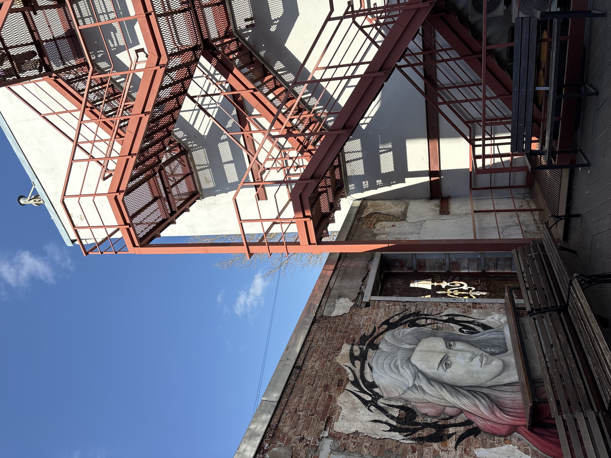 The scene is an urban outdoor area located in Saint Petersburg, Russia, observed under a clear blue sky with minimal white clouds. No individuals are present. The foreground is covered by a wooden deck.
On the left, a weathered brick wall, partially obscured by peeling light-colored plaster, features a large graffiti mural depicting a woman's face with long, light-colored hair, framed by a dark, thorny, or spiky motif. A red garment is visible around the lower part of the mural. To the immediate right of the mural, a dark, dilapidated wooden door is set into the wall, reflecting an ornate, gold-toned pattern. Two dark wooden benches with metal frames are situated on the deck in front of the mural, one incorporating a narrow wooden table.
On the right, a multi-story building with a smooth, light-colored facade is prominent. A large, external fire escape, constructed from red-painted metal with expanded mesh stairs and landings, ascends the building in a zig-zag pattern. Direct sunlight creates distinct, geometric shadows of the fire escape structure on the building's facade. A small, metallic, cylindrical object is visible atop the building against the sky. A third dark wooden bench with a metal frame is positioned on the wooden deck beneath the fire escape, with multiple outdoor units, likely air conditioning, visible behind it.