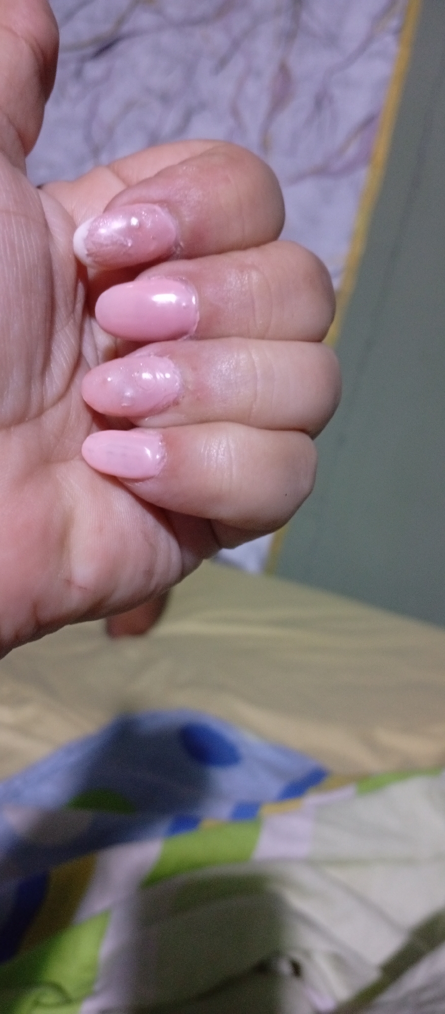 A close-up shot focuses on a person's hand with polished fingernails. The fingernails are painted a pale pink with a glossy finish, with the thumb's nail featuring a white tip. The hand appears slightly wrinkled, suggesting an older individual. The background includes a bed with patterned sheets in shades of blue, green, and white. The presence of sheets suggests the location is indoors, possibly a bedroom or similar resting area. There are no clear indicators to specify the exact location of Juan Griego, Venezuela. The lighting is soft and diffused, with no distinct shadows, providing a uniform illumination. The image primarily captures a hand with a manicure against a blurred backdrop.