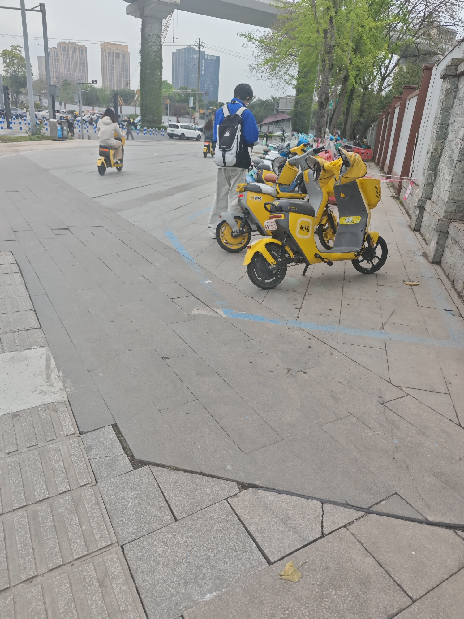 The scene is a street in Chongqing High-tech Zone, China. A group of yellow electric scooters are parked along a wall. A person in a blue shirt and backpack stands near the scooters. Another person rides a similar scooter on the street. Buildings and an elevated structure are visible in the background.