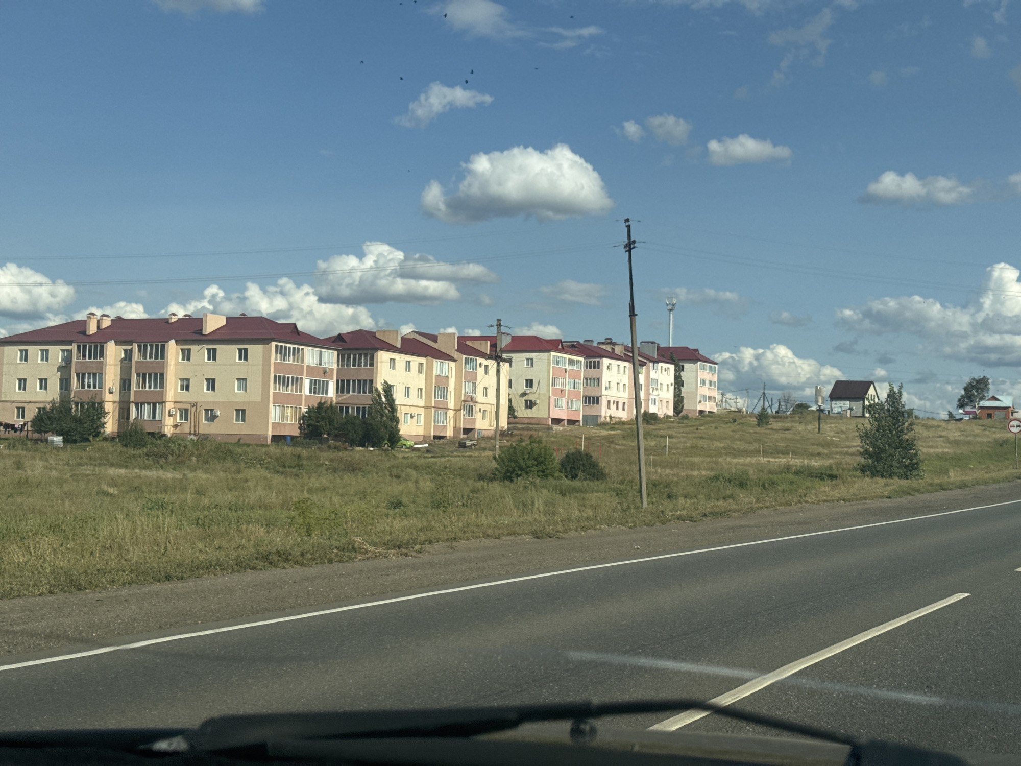 The image depicts a row of multi-story apartment buildings with red roofs situated on a slight incline, overlooking a road. The buildings appear to be constructed with a light-colored facade and have numerous windows and balconies. A grassy field with scattered trees separates the buildings from the road, which is asphalt with white lane markings. In the background, a cloudy blue sky is visible with some birds flying. There are also utility poles and wires present. To the right of the apartment buildings, there is a smaller house with a dark roof, and further in the distance, another building with a red roof. A "no vehicles" sign is visible on the right side of the road. The scene suggests a semi-urban or suburban environment in Novosergiyevka, Russia, likely during the daytime under fair weather conditions. No people are clearly discernible in the image.
