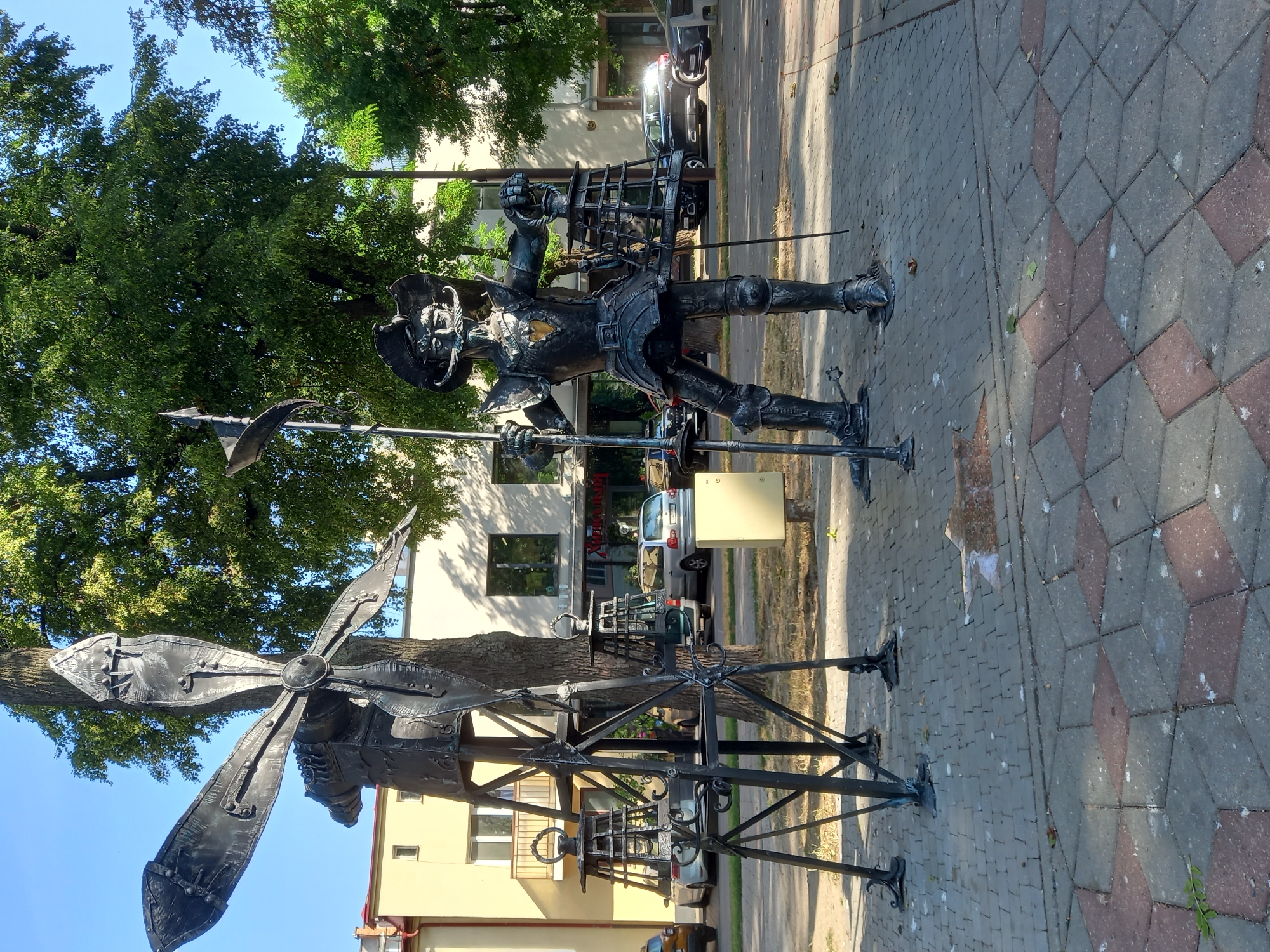 In Brest, Belarus, a metal sculpture depicting a knight stands on a paved walkway. The knight is dressed in armor and wears a hat with a wide brim and a prominent mustache. He holds a long spear in his left hand and a lantern in his right. To the left of the knight is another metal sculpture resembling a windmill with large blades. The background shows trees, a yellow building with balconies, and parked cars. The walkway is made of interlocking paving stones, with a star-shaped pattern of red and grey stones in the foreground. The scene appears to be outdoors on a sunny day.