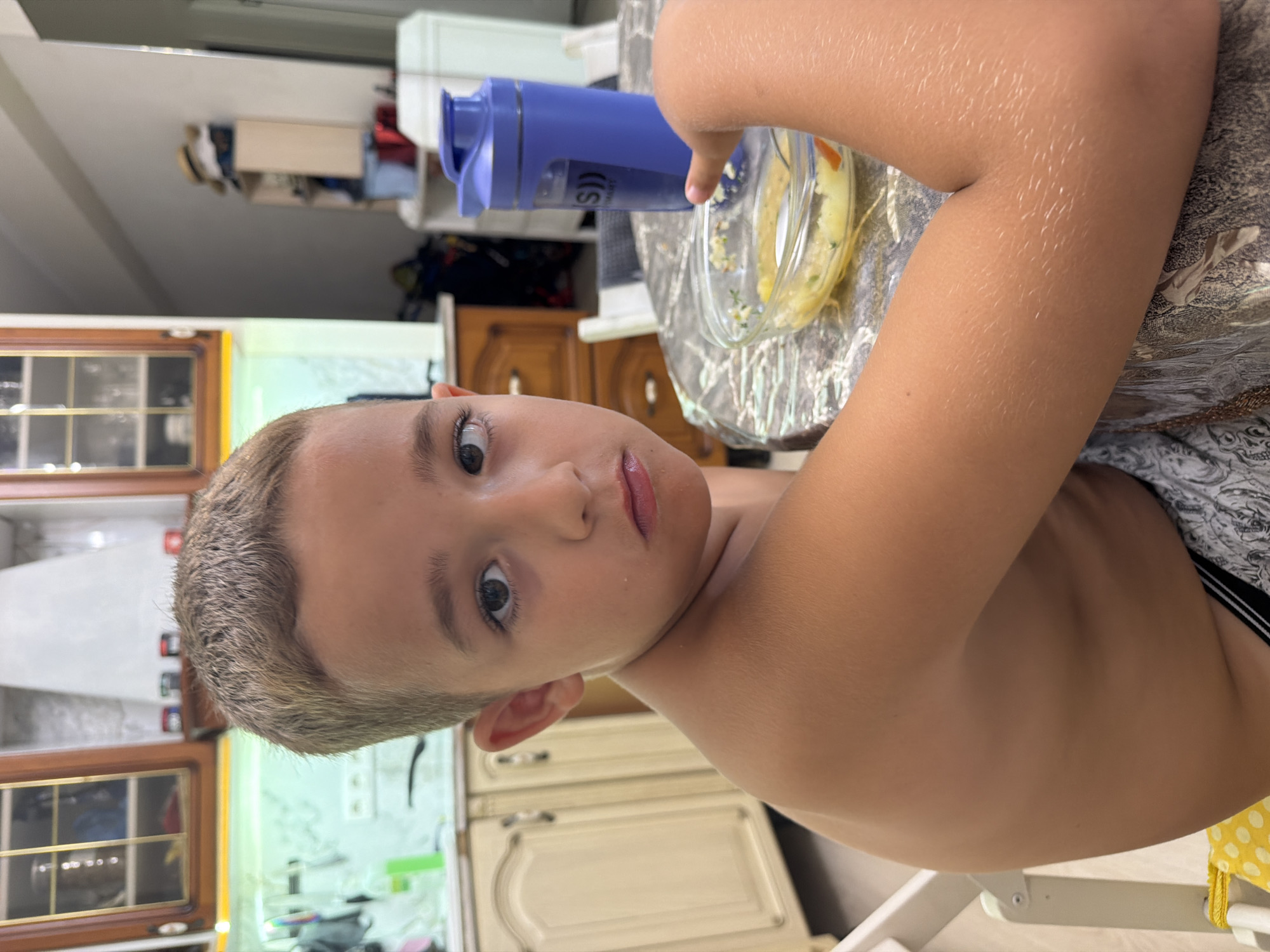 A boy, appearing to be around 5-7 years old, is shown from a close-up, slightly angled perspective. He has short, light brown hair and is shirtless. His facial features are prominent, with large, dark eyes and slightly parted lips. He is looking towards the camera with a neutral or slightly curious expression.
The boy is seated at a table with a textured, possibly wooden surface covered by a patterned placemat. To his right, a clear glass bowl contains a light-colored food, possibly a meal or dessert, with small pieces of orange and green visible within it. Next to the bowl is a blue, opaque water bottle with a black lid and a silver band around its neck. Text is visible on the water bottle, though it is partially obscured and difficult to read clearly.
The background suggests an indoor setting, likely a kitchen or dining area. Visible elements include kitchen cabinets with wooden doors and glass panes, a light-colored countertop, and what appears to be a refrigerator or another tall cabinet. The overall lighting suggests it is daytime, with no indication of specific weather conditions. The image appears to be taken in Sochi, Russia, as per the location context. The boy's skin has a slightly rosy hue, and there are visible goosebumps on his arm, suggesting a cool environment or a reaction to something.