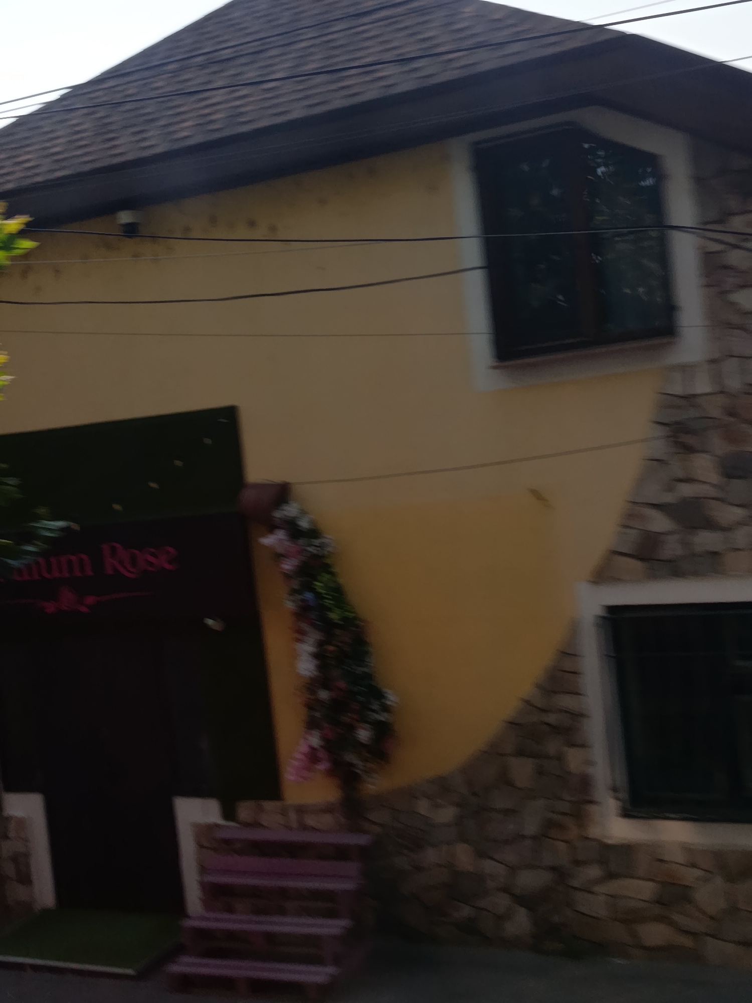 The image shows a portion of a building with a brown shingled roof. The building has a yellow facade with a stone foundation. There is a dark-colored sign with pink lettering that reads "mum Rose" on the left side of the image, partially obscured by a dark object. To the right of the sign, there is a decorative garland with flowers and foliage. Two windows are visible on the building; one is a large, arched window on the upper floor, and another is a smaller, rectangular window on the lower floor. Power lines are visible against the sky. The scene appears to be outdoors, possibly in a residential or commercial area. The image is of a building in Cahul, Moldova.