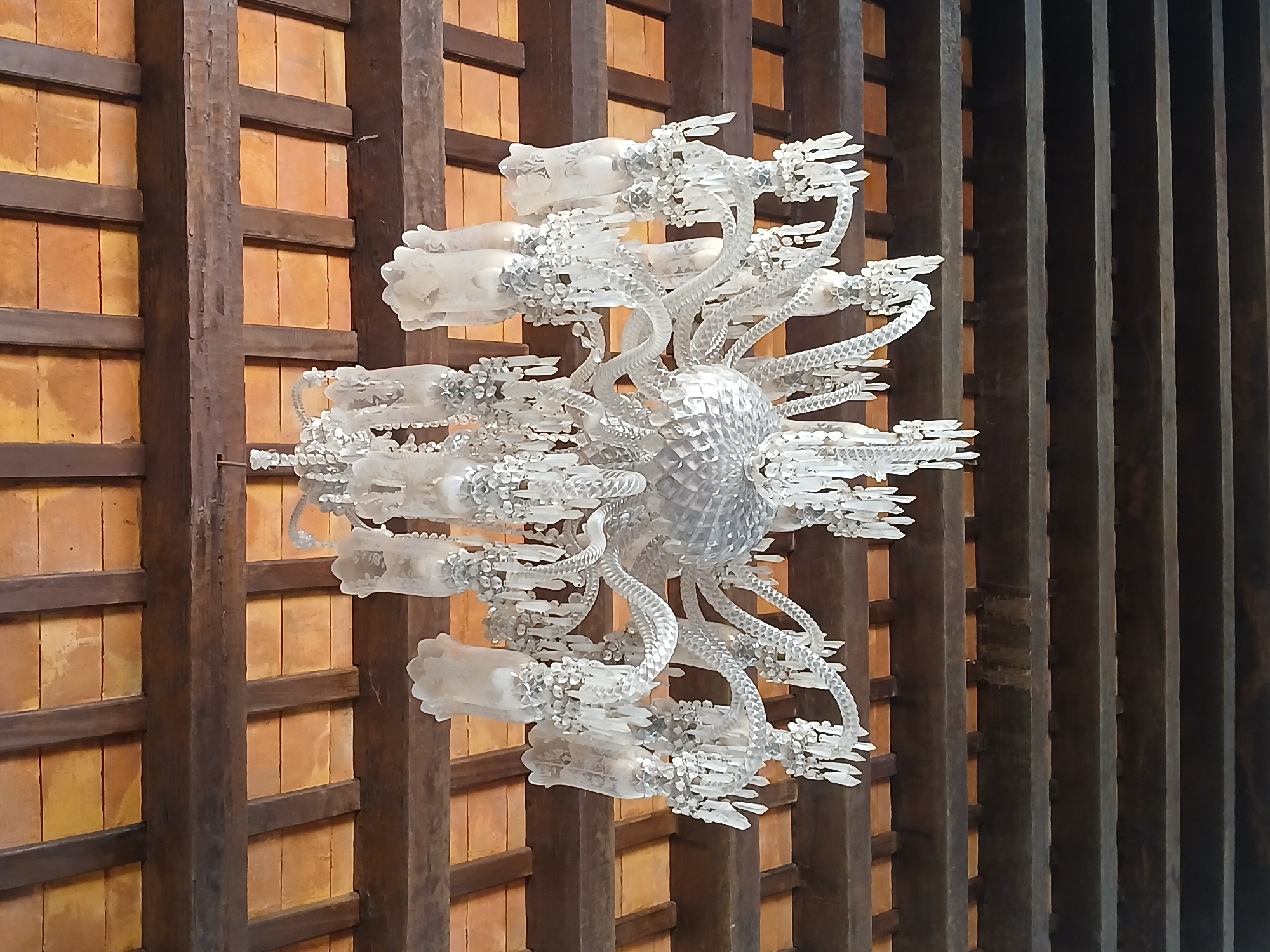 The image shows a decorative chandelier-like object attached to a wooden ceiling structure. The ceiling is comprised of wooden beams arranged in a grid pattern, with tan rectangular panels between them. The chandelier is made of translucent, possibly glass or plastic, materials and has a complex, organic design with a central, textured sphere and numerous branching arms. The specific location in Ciudad Bolivar, Venezuela is not clear from the image.