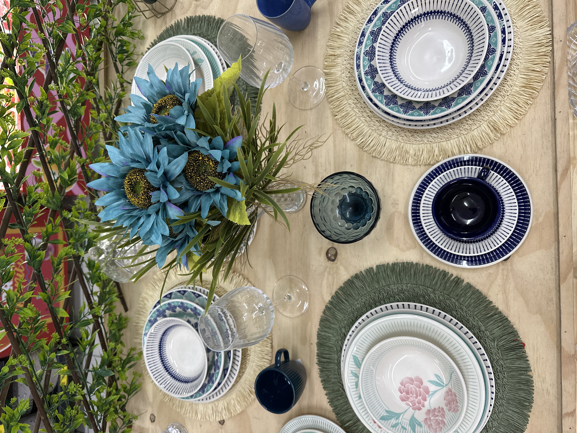 The image shows a table set for a meal, likely in Florianópolis, Brazil, with a rustic wooden table as the surface. The table is adorned with placemats, dishes, and glassware, with a floral arrangement serving as a centerpiece. The placemats are of two types: one set is made of woven straw with a fringed edge, and another is a dark green woven circular mat with a fringed edge.
On the straw placemats, there are stacks of white plates with blue and teal geometric patterns. Each stack includes dinner plates, smaller plates, and bowls. On one of these stacks, a dark blue teacup is placed. On the other straw placemat, a single teacup is present.
The green placemats feature white plates with pink floral designs. One placemat has a stack of these plates, and a dark blue teacup sits on top of it.
Glassware includes clear wine glasses with textured patterns, and a dark blue patterned glass bowl is placed on the table. A bouquet of artificial blue sunflowers with yellow centers and green foliage is arranged in a clear glass vase at the center of the table.
Two dark blue ceramic mugs are also visible, one near a wine glass on the straw placemat and another on the right side of the image. The background features greenery and what appears to be a decorative screen or fence, possibly indicating an outdoor or semi-outdoor setting, or a setting designed to mimic one. The lighting suggests it is daytime.