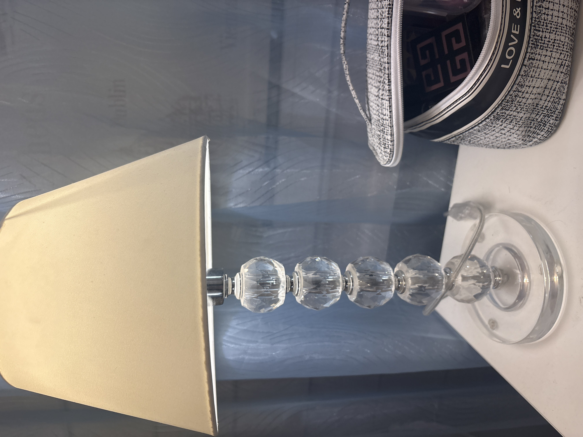 A close-up, slightly angled shot shows a bedside table with a lamp and a makeup bag. The lamp has a cream-colored shade and a base made of stacked crystal spheres. To the right, a patterned makeup bag is open, revealing makeup items inside. The background features a translucent blue curtain and a wall with faint lettering. The scene is indoors, likely in a residential setting, with no people visible.