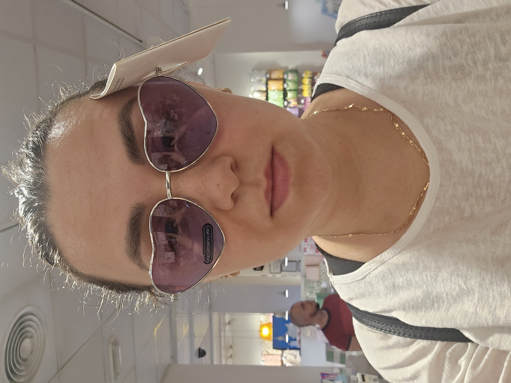 A close-up, head-on, selfie-style shot features a woman wearing large, purple, heart-shaped sunglasses with a price tag attached to the frame. The sunglasses are reflective, showing a blurred image of a store interior with shelves and other shoppers. The woman has her hair pulled back, with several strands of hair escaping around her forehead and temples. She is wearing a white t-shirt and a delicate gold chain necklace. The background is softly blurred, suggesting an indoor retail environment, possibly a clothing or accessories store. There is no visible indication of the city of Jaén, Spain.