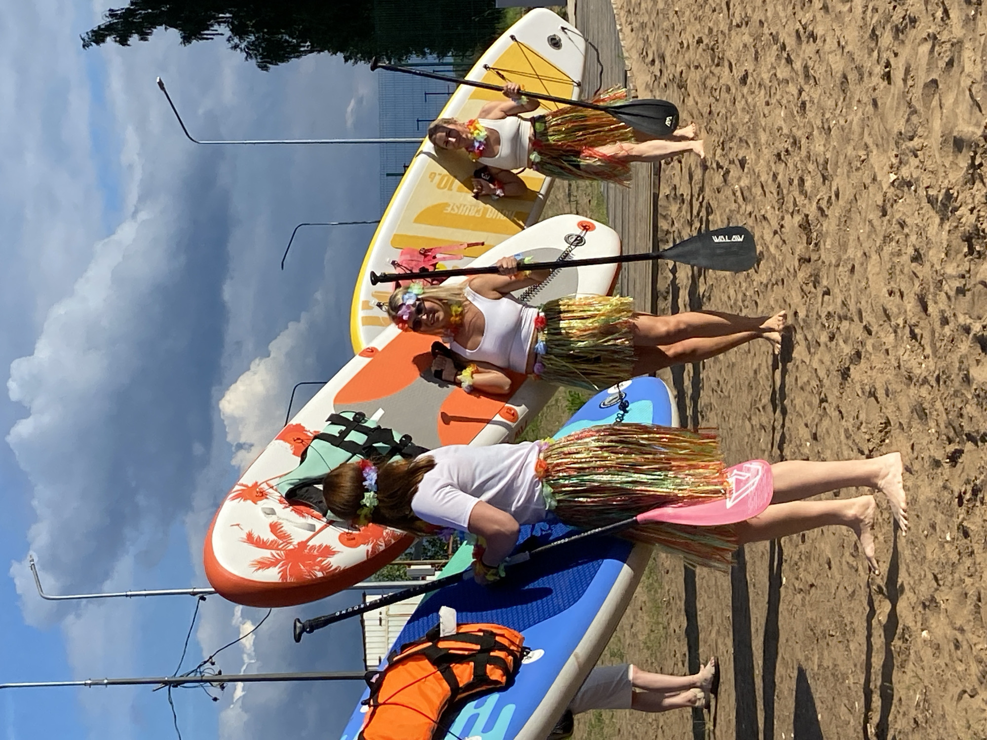 The image shows three women dressed in white tops and colorful hula skirts, posing with paddleboards on a sandy beach. They are wearing flower leis around their necks and some have flower crowns in their hair, suggesting a festive or themed event. Two of the women are holding paddles. The paddleboards are stacked and arranged in a way that suggests they are about to engage in paddleboarding or have just finished.
The scene is outdoors, with a cloudy sky and bright sunlight, indicating it is daytime. The background shows a grassy area and some fencing, with a body of water potentially in the distance, though not clearly visible. The sand and the presence of paddleboards suggest a location near a lake, river, or sea. The presence of streetlights and a wooden deck also points to a recreational area.
One paddleboard has the text "VALAV" visible on the paddle. Another paddleboard has "PROCRUISE" printed on it. The overall mood appears to be lighthearted and fun, with the women smiling and posing for the camera. The event seems to be a recreational activity, possibly a bachelorette party, a team-building event, or just a fun outing with friends. The time of day appears to be afternoon, given the sunlight and shadows.