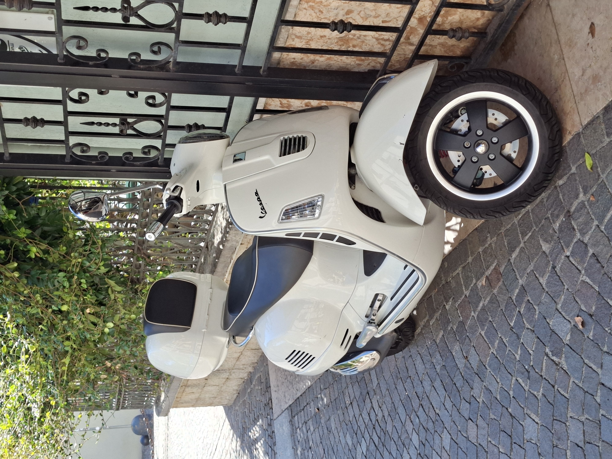A white Vespa scooter with a black seat and black top box is parked on a cobblestone sidewalk next to a black metal gate. The scooter has a silver-colored trim around the handlebars and mirrors. The gate has ornate, wrought-iron scrollwork. Behind the gate, a building with a light-colored facade is visible, and further back, green foliage from a bush or tree hangs over the fence. The scene is lit by sunlight, casting shadows on the ground. The scooter is the primary subject, positioned at an angle in the foreground. The setting appears to be an outdoor urban environment in the city of Peschiera del Garda, Italy, suggested by the architectural style of the gate and building, and the cobblestone paving. There are no people visible in the image.
