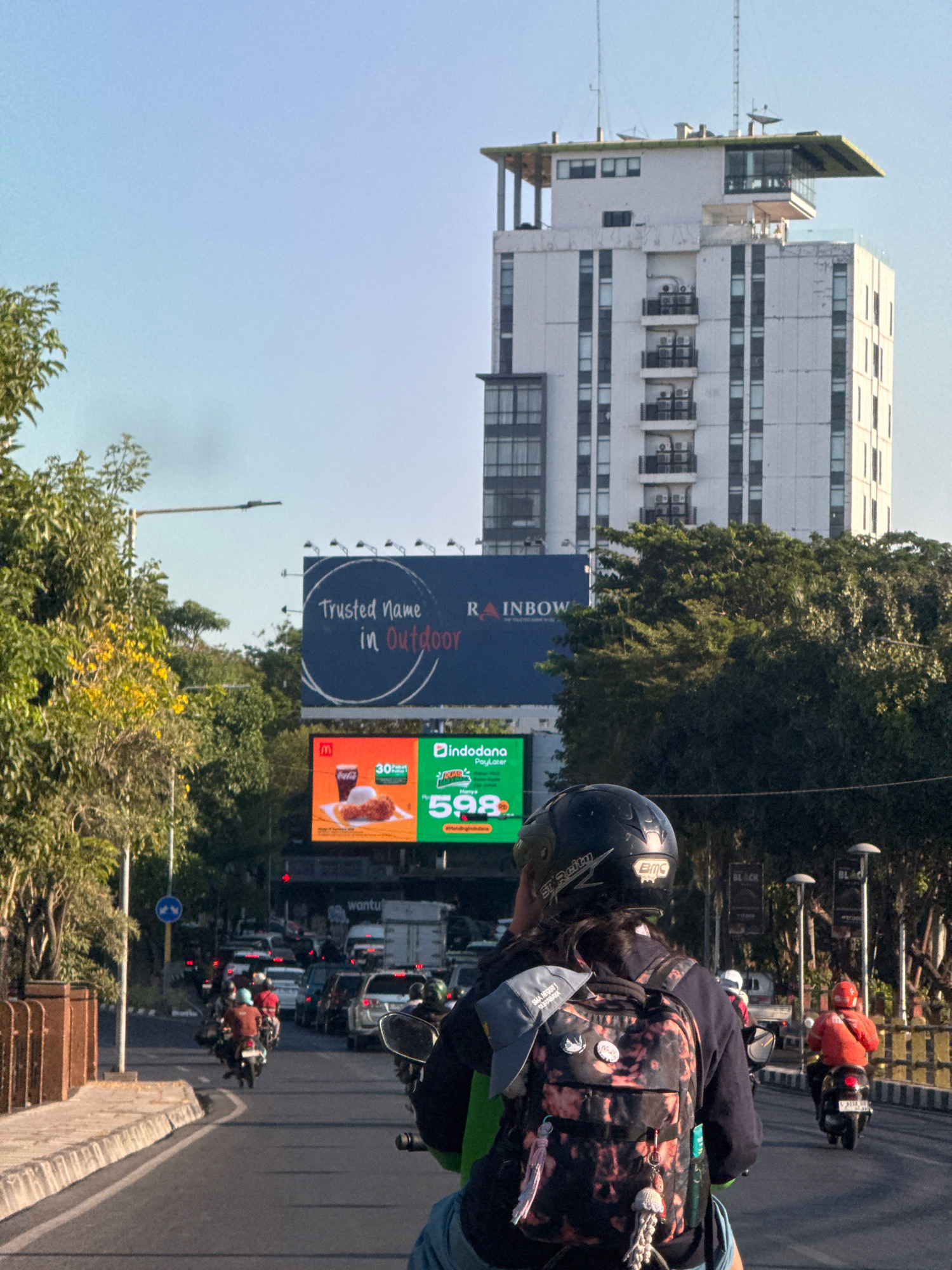 A person wearing a helmet and backpack rides a motorcycle on a busy street in Surabaya, Indonesia. The rider is in the foreground, with the motorcycle's handlebars and a portion of their body visible. The backpack is adorned with patches and a keychain. In the background, a tall, modern building with balconies stands against a clear sky. Billboards are also visible; one displays the text "Trusted Name in Outdoor," and another promotes a food item with the price "598." The street is filled with other motorcycles and cars, indicating moderate traffic. Trees line the sides of the road, and streetlights and traffic signals are present. The overall scene captures a snapshot of daily life in an urban environment.