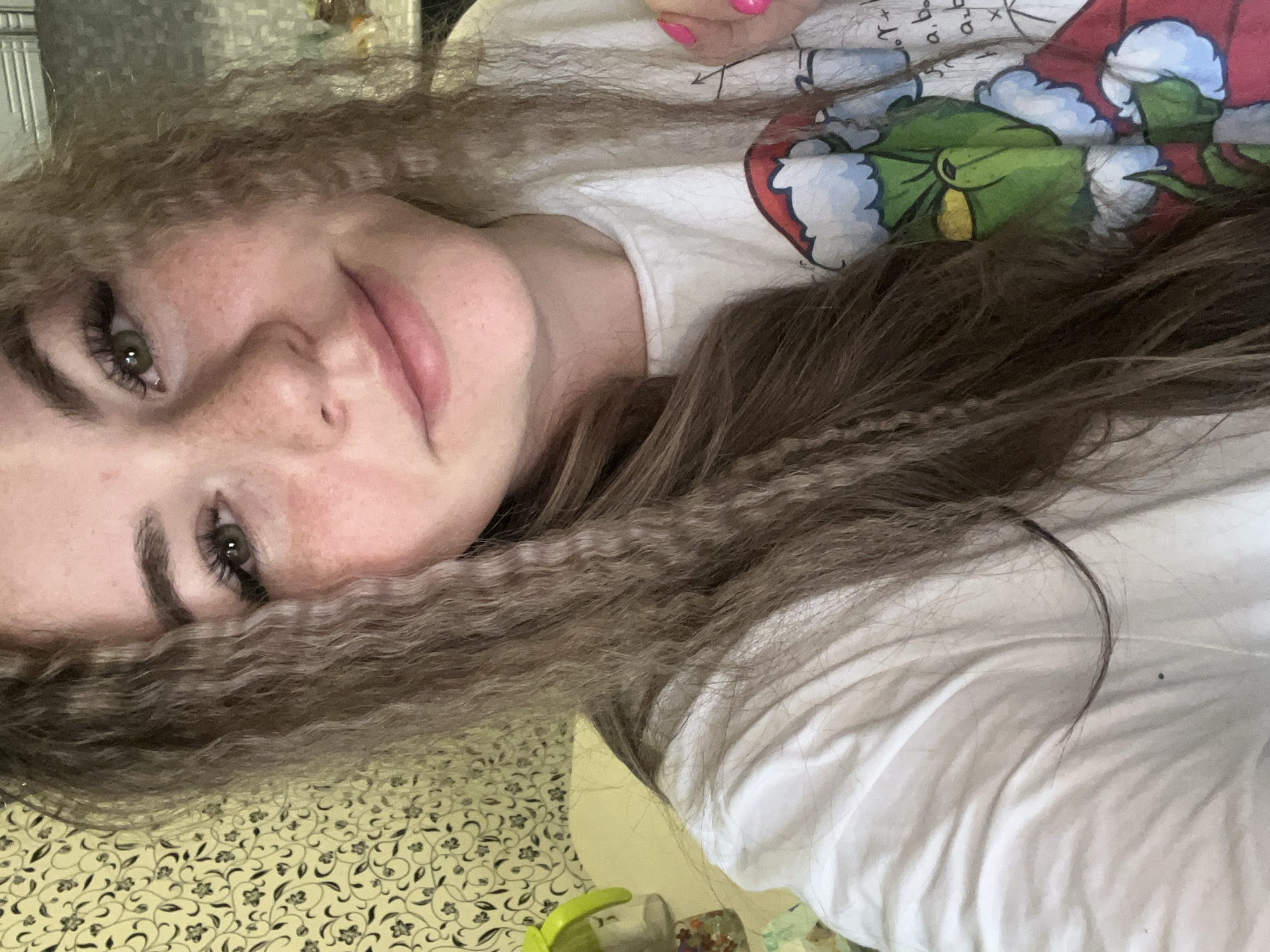 A young woman with brown, crimped hair is seen in a selfie. She has fair skin with freckles and green eyes. She is wearing a white t-shirt with a graphic of The Grinch. Her fingernails are painted pink. The background shows a patterned surface and part of what appears to be a white surface. It is not possible to determine the location in Chaikovsky, Russia, from the provided image.