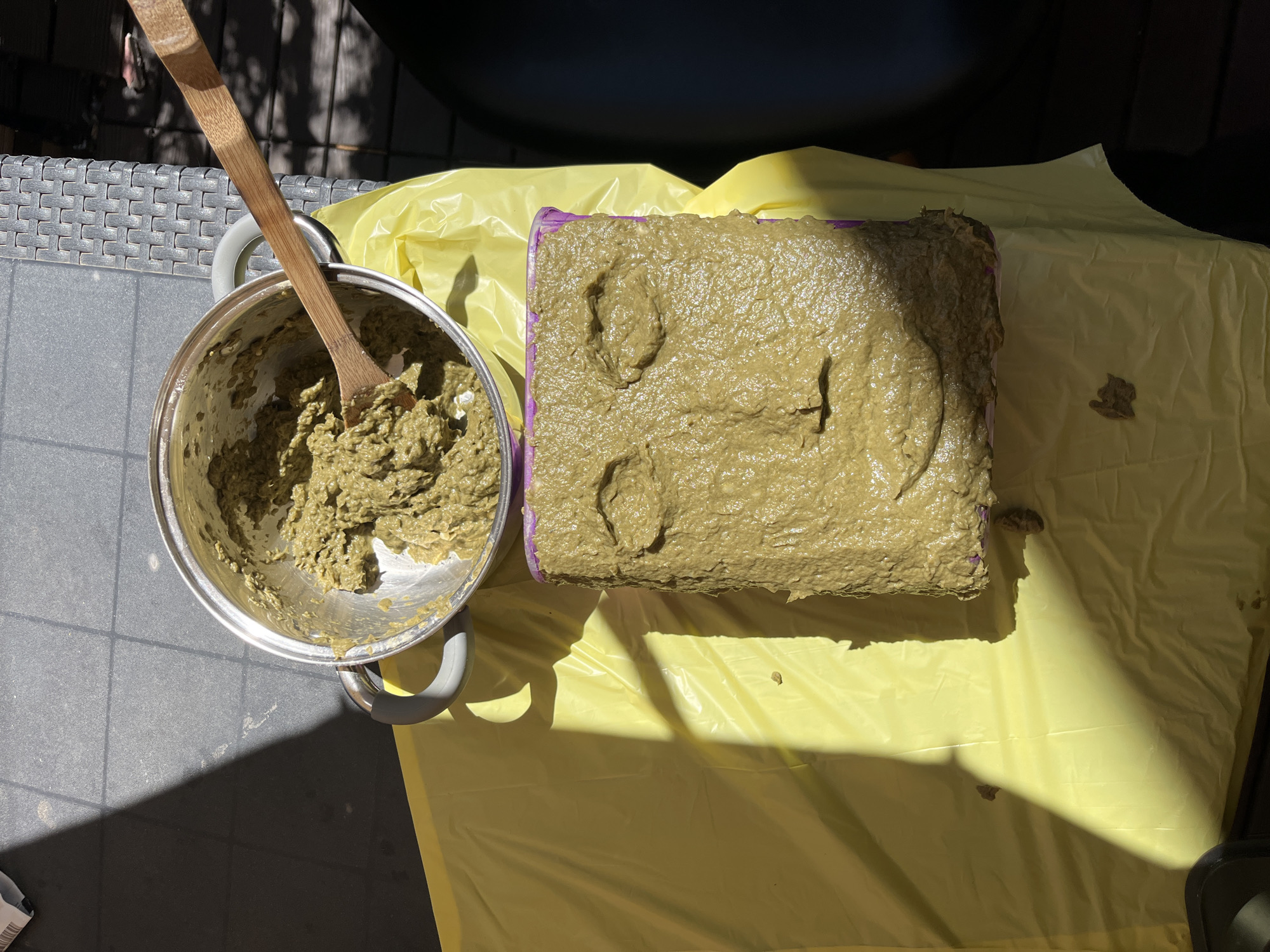 An overhead shot captures an outdoor scene featuring a pot containing a thick, greenish-brown paste, stirred with a wooden spoon. Adjacent to the pot, a rectangular container or mold is filled with the same pasty substance, displaying several deep indentations or impressions on its surface, resembling abstract footprints or shapes. Both items rest on a bright yellow plastic sheet or tarp, which is spread over a grey tiled floor, likely a patio or balcony.
The scene is brightly lit by strong sunlight, casting prominent, dark shadows across the tiled surface and the yellow sheet, indicating it is daytime. In the background, part of a grey, textured outdoor furniture piece, possibly rattan, is visible, along with a dark, indistinct object. There are no people visible, and no discernible text in the image. The setup suggests a crafting activity, possibly involving molding or making impressions in a clay-like or plaster-like material.