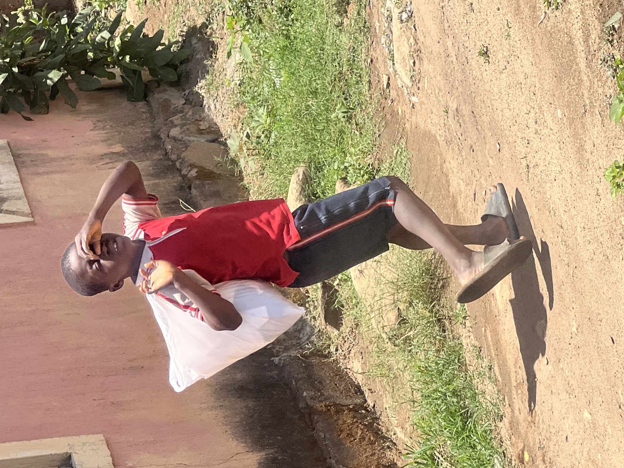 The image shows a young Black male, likely a boy or teenager, standing outdoors on a sunlit dirt path. He is wearing a red short-sleeved shirt with white accents, dark blue shorts with red and white side stripes, and grey slip-on sandals. He holds a large white bag, possibly plastic or cloth, against his left side. His right hand is raised to his forehead, shielding his eyes from the bright sun, indicating it is likely midday or early afternoon with clear, sunny weather.
The scene features a light red or pink building wall and a lower concrete or stone structure on the left side of the frame. To the right and in the background, there is green vegetation, including grass and some larger leafy plants, bordering the dry and dusty path. Strong shadows are cast on the ground, further emphasizing the intense sunlight. The setting appears to be a casual, everyday outdoor environment in a warm climate, consistent with the location context of Gura Top, Nigeria. There is no visible text in the image.
