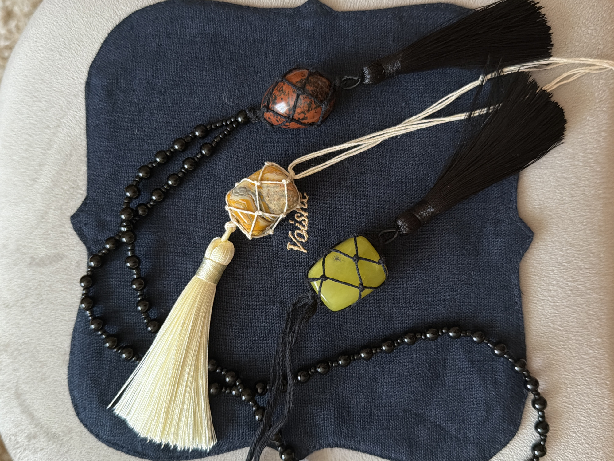 The image shows three beaded necklaces with decorative pendants, lying on a dark blue fabric surface with a small golden logo "Voish". The fabric is resting on a light-colored, possibly beige or light gray, plush surface.
The necklaces consist of black beads. Each has a different colored stone enclosed in a macrame-style net with a tassel attached. One necklace has a red and black stone with a black tassel, another has a beige and yellow stone with an off-white tassel, and the third has a yellow-green stone with a black tassel.
The photo is taken in a neutral indoor lighting environment.
There is no evidence to place the location of the photo in Khimki, Russia. The image can not be described as having any city context.