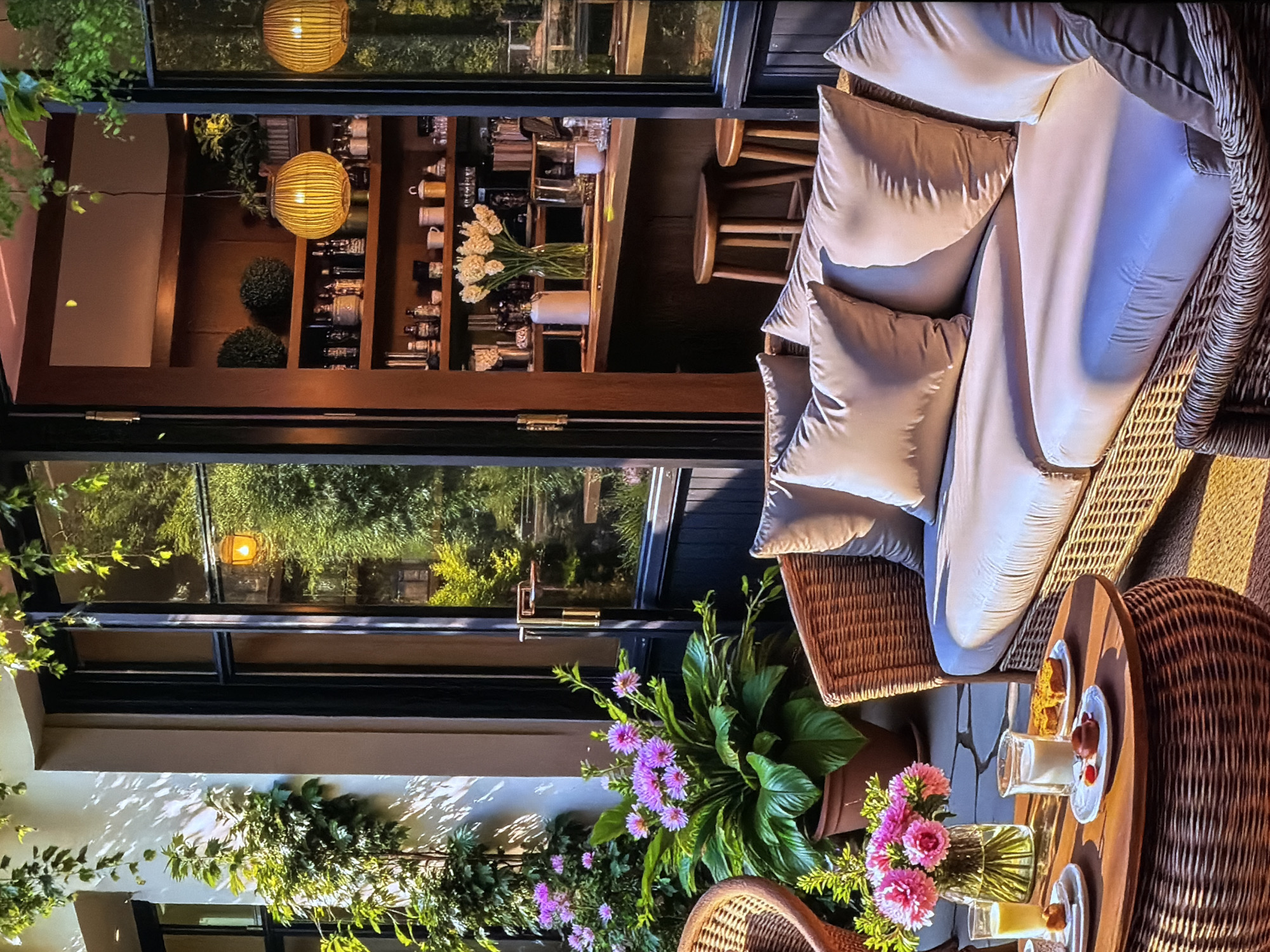The image displays a warm and inviting outdoor seating area, possibly a patio or balcony, connected to an indoor space through large glass windows or doors. The setting suggests a cozy café, restaurant, or a well-appointed home in Sofiivska Borshchahivka, Ukraine.
In the foreground, a comfortable, light-colored wicker or rattan sofa is furnished with several plush light gray cushions and pillows. Adjacent to it, a matching round wicker table is set for a snack or dessert, holding two small white plates with portions of cake or pastry, and two glasses that appear to contain milk or a light beverage. A clear glass vase filled with vibrant pink chrysanthemums or similar flowers adds a decorative touch to the table. Lush potted plants, including one with broad green leaves and purple daisy-like flowers, and climbing green foliage on a wall, enhance the natural aesthetic of the outdoor space.
Through the glass partition, an indoor area is visible, resembling a bar or serving counter. It features a wooden shelving unit stocked with various bottles, mugs, and glassware. A vase of white flowers (possibly tulips) sits on the counter. Two simple wooden bar stools are positioned near the counter. Two spherical, woven light fixtures hang from the ceiling, casting a warm yellow glow within the interior.
The arrangement implies a relaxed activity such as enjoying a dessert or beverage. The time of day appears to be late afternoon or early evening, indicated by the warm, directional sunlight creating distinct shadows on the outdoor wall and the glowing interior lights, contributing to a "golden hour" ambiance. The weather seems pleasant, ideal for outdoor relaxation. The overall atmosphere evokes comfort, tranquility, and hospitality. There is no visible text in the image.