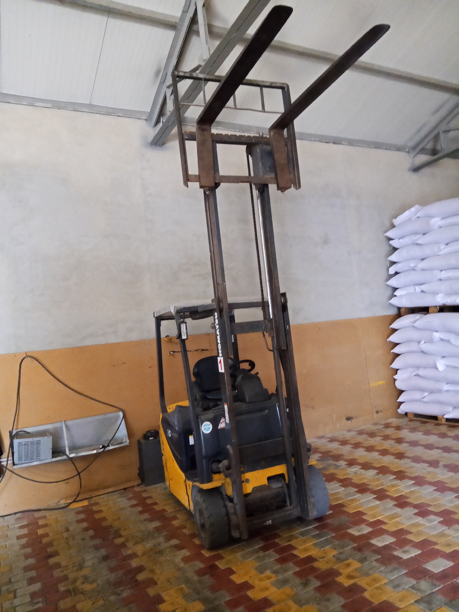The image depicts the interior of a warehouse. A yellow Komatsu forklift is positioned in the center of the frame, its forks raised high. To the right of the forklift, stacks of white sacks are piled on wooden pallets. The floor is paved with interlocking blocks in shades of red, yellow, and grey. The walls are painted a light, neutral color, and the ceiling has exposed metal framing. In the background, near the left wall, there is a metal shelf with a piece of electrical equipment. The overall scene suggests a storage or distribution facility. The image does not contain any people or specific indicators of its location within Ceadîr-Lunga, Moldova.