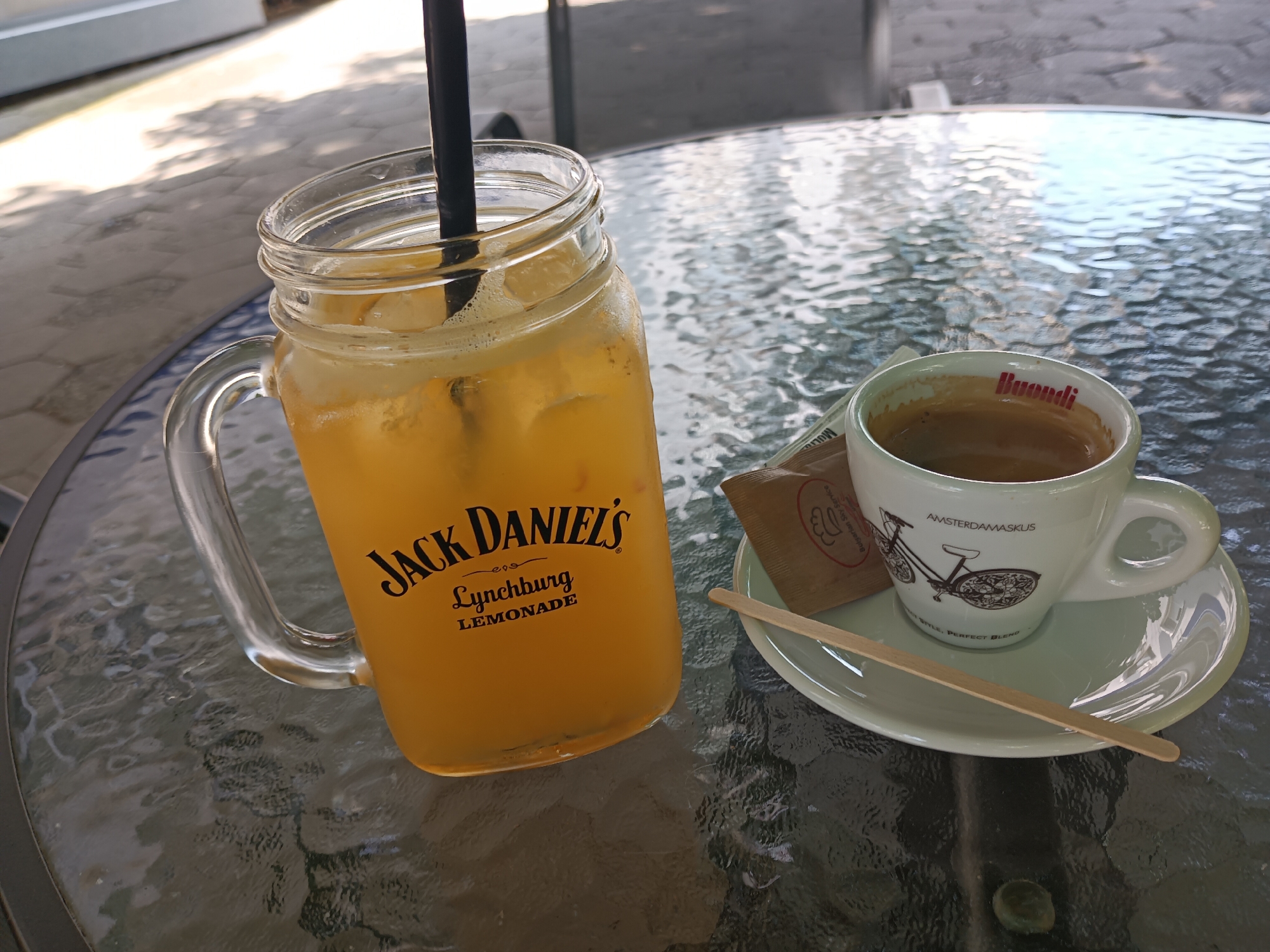 The image shows a glass mug with "Jack Daniel's Lynchburg Lemonade" printed on it, filled with a yellow beverage and ice, and a black straw. Beside the mug is a white cup of coffee on a saucer, with a packet of sugar and a wooden stirrer. The items are placed on a glass table, which is outdoors, with dappled sunlight suggesting it is daytime. The table is set on an outdoor paved area, possibly a cafe or restaurant patio, with some blurred background elements that could indicate urban surroundings. The scene is forensic and matter-of-fact; there are no people or interactions depicted, only the arranged objects and environment. The request for a specific location within Burgas, Bulgaria, cannot be fulfilled as there are no discernible landmarks or contextual clues within the image to confirm this location.