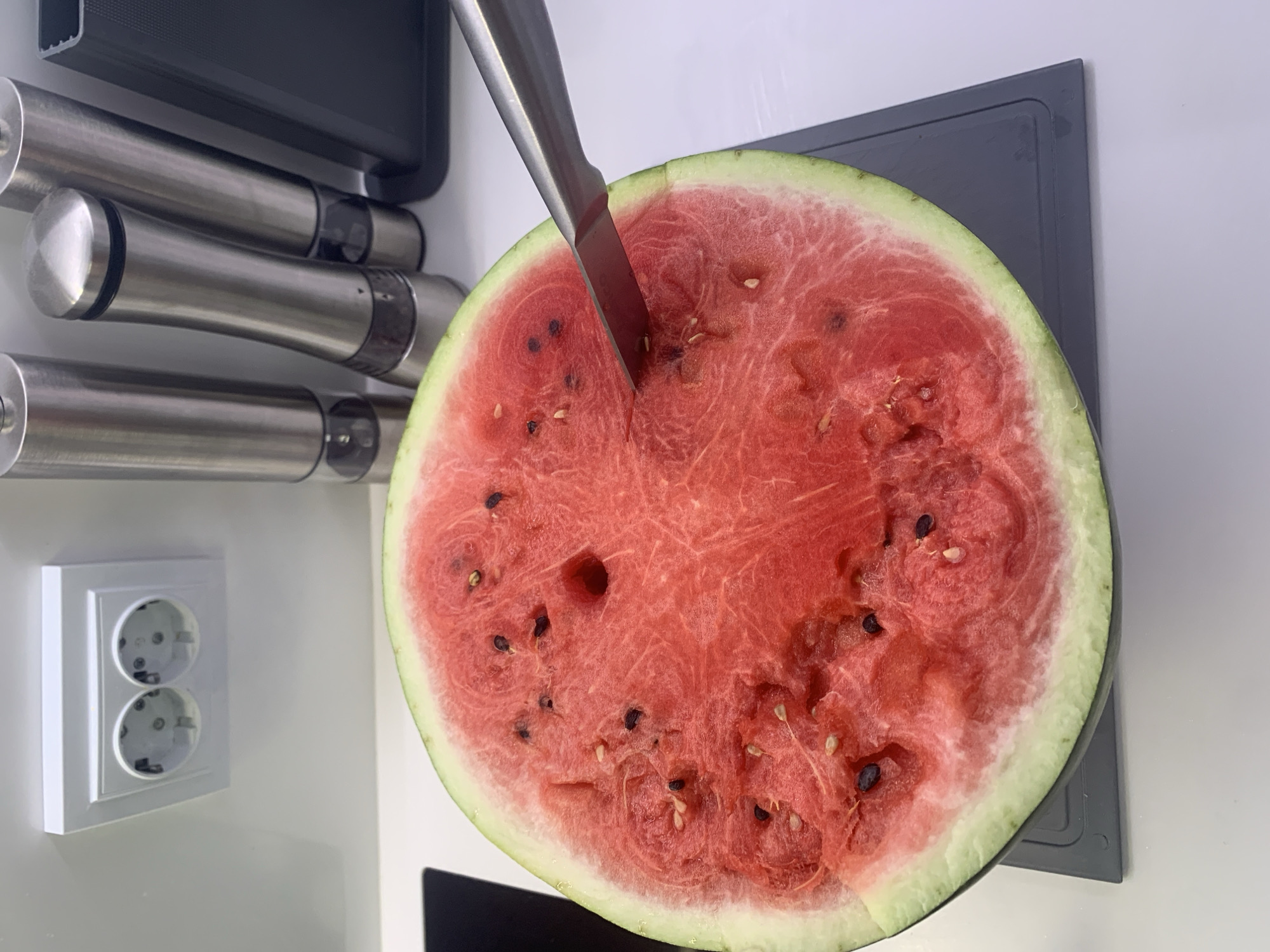 A close-up, eye-level shot shows the interior of a halved watermelon on a gray cutting board. The watermelon flesh is a vibrant red, speckled with numerous black seeds. A silver knife is inserted into the watermelon, angled downwards. In the background, out of focus, are two stainless steel salt and pepper grinders and a white electrical outlet with two circular sockets. The overall setting appears to be a kitchen. The image was captured in Bugry, Russia.