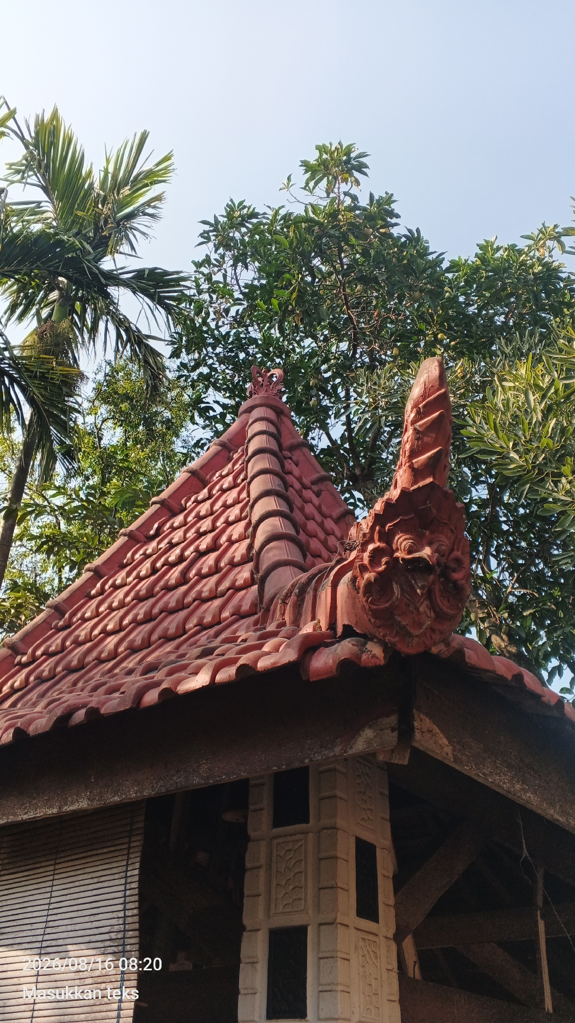 The image shows the roof of a building with red clay tiles, adorned with decorative carvings. A prominent carved feature, resembling a mythical creature or dragon, is visible on the right side of the roof ridge. Green foliage from trees surrounds the structure. The sky is clear and blue. The timestamp "2026/08/16 08:20" and the text "Masukkan teks" are present in the lower left corner, indicating the image was taken at that time and date in Kendalpayak, Indonesia, and is likely a photograph taken with a mobile device.
