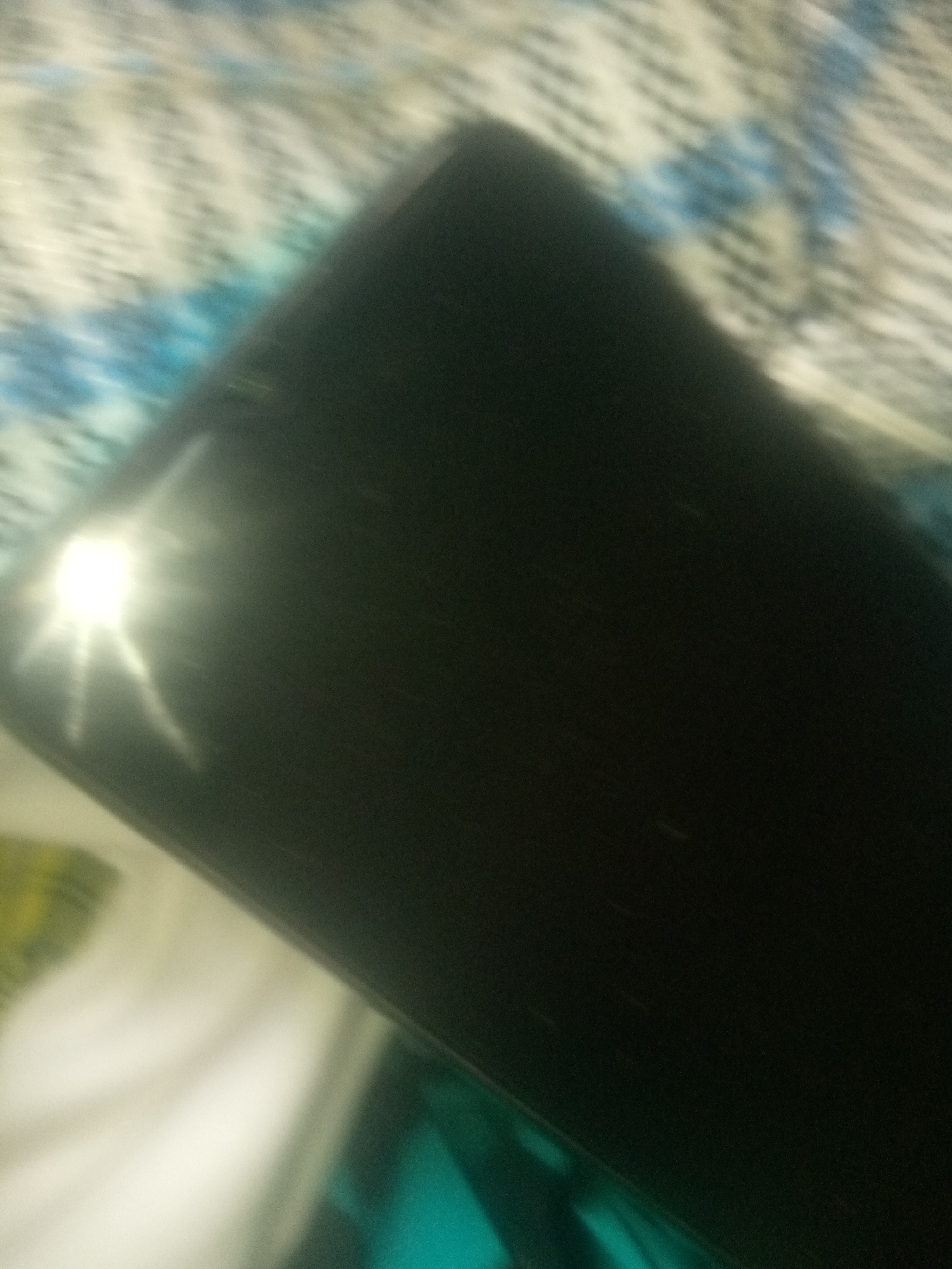 A black smartphone is resting on a patterned surface. The phone's screen is dark, with a bright light source creating a lens flare effect on the left side. The patterned surface appears to be a fabric with abstract blue and white designs. The image is blurry and taken from a slightly elevated angle.